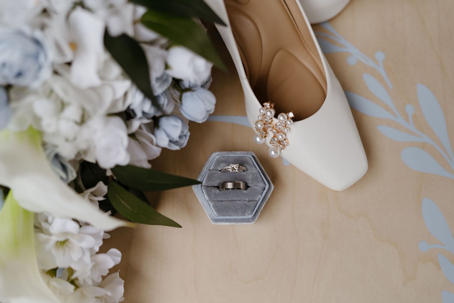 Wedding rings in a gray velvet box beside white bridal heels and a bouquet of blue and white flowers.