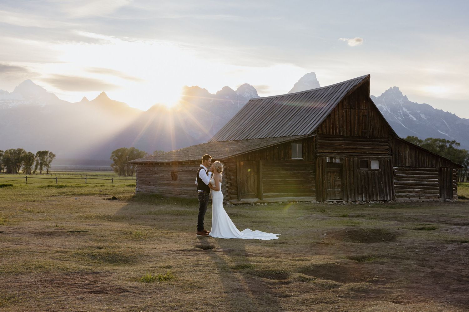 Wedding couple embracing at sunset in front of historic Mormon Row barn with Grand Teton mountains.