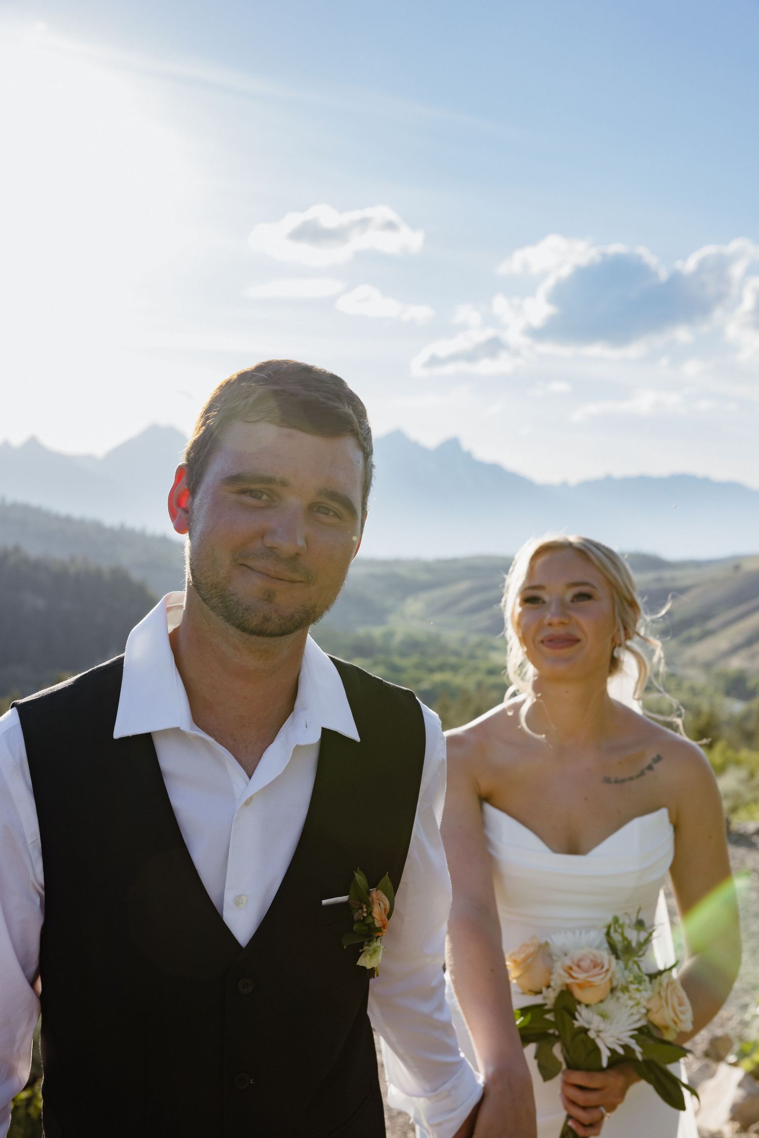 Groom in black vest smiles at camera as bride holds floral bouquet with sunlit Teton mountains behind them.