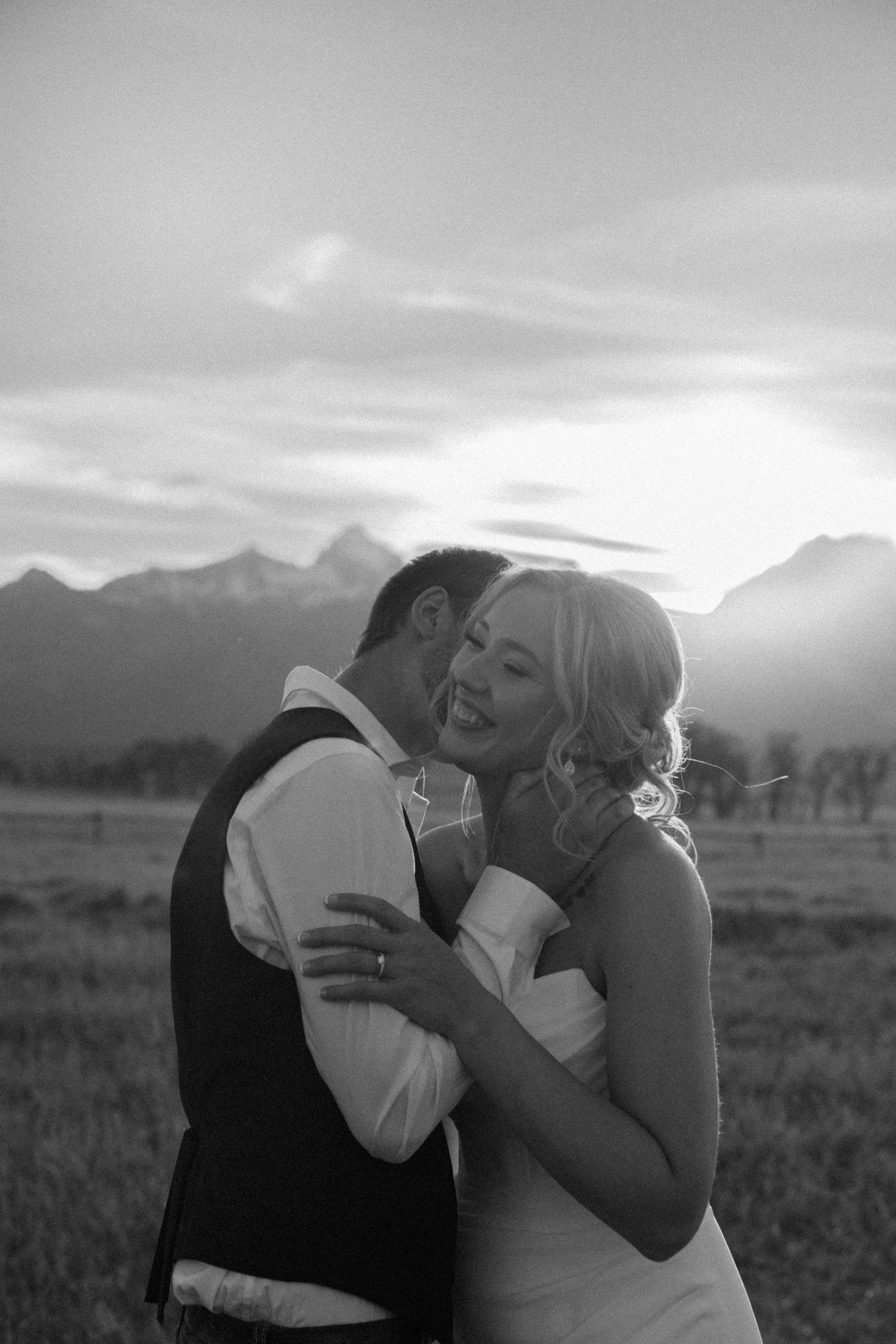 Black and white photo of laughing bride being kissed by groom with mountain landscape at sunset.