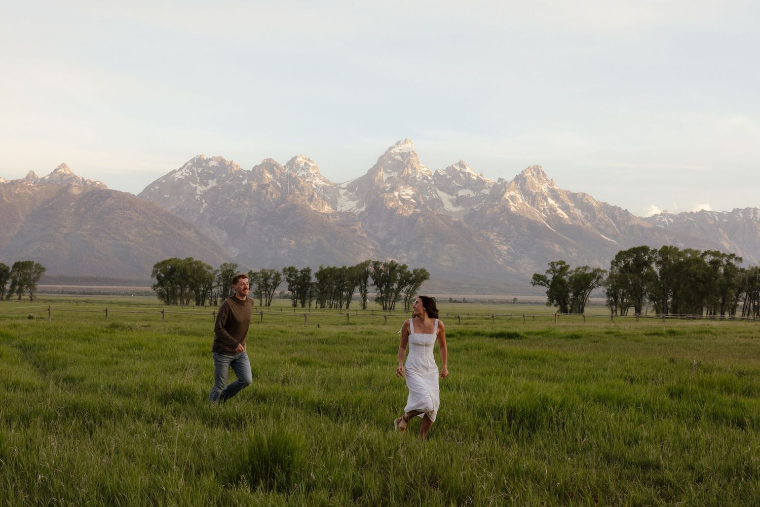 Couple walking through green meadow with stunning Grand Teton mountain range in the background at dusk.