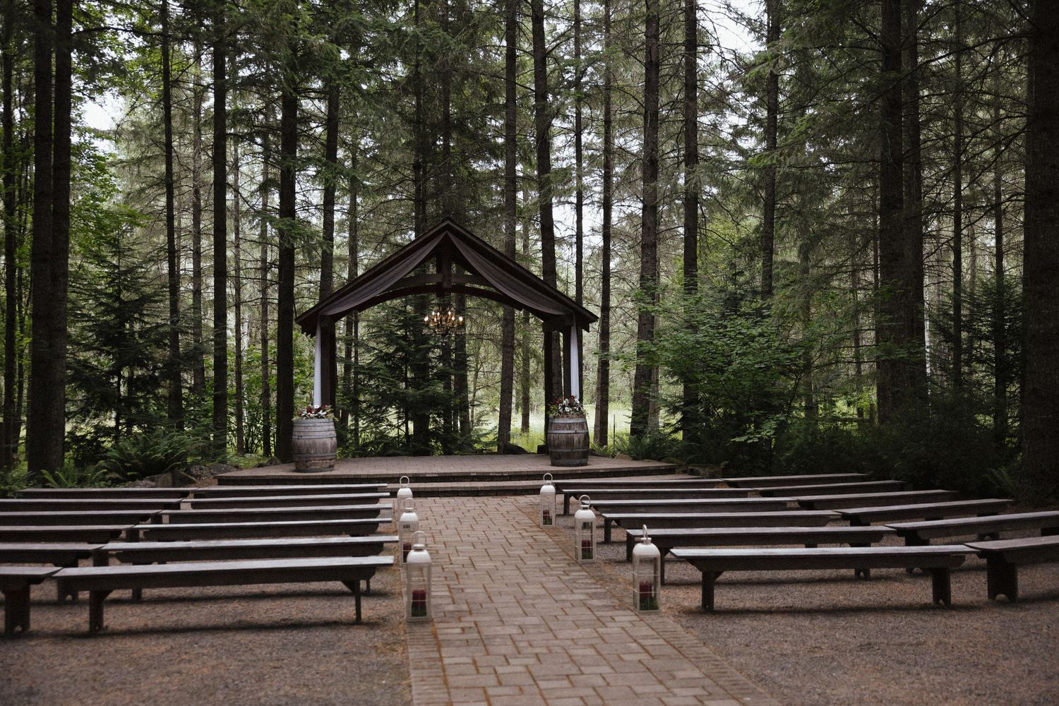 Empty outdoor forest wedding ceremony venue with wooden benches, brick aisle, and rustic wooden arch among tall pine trees.