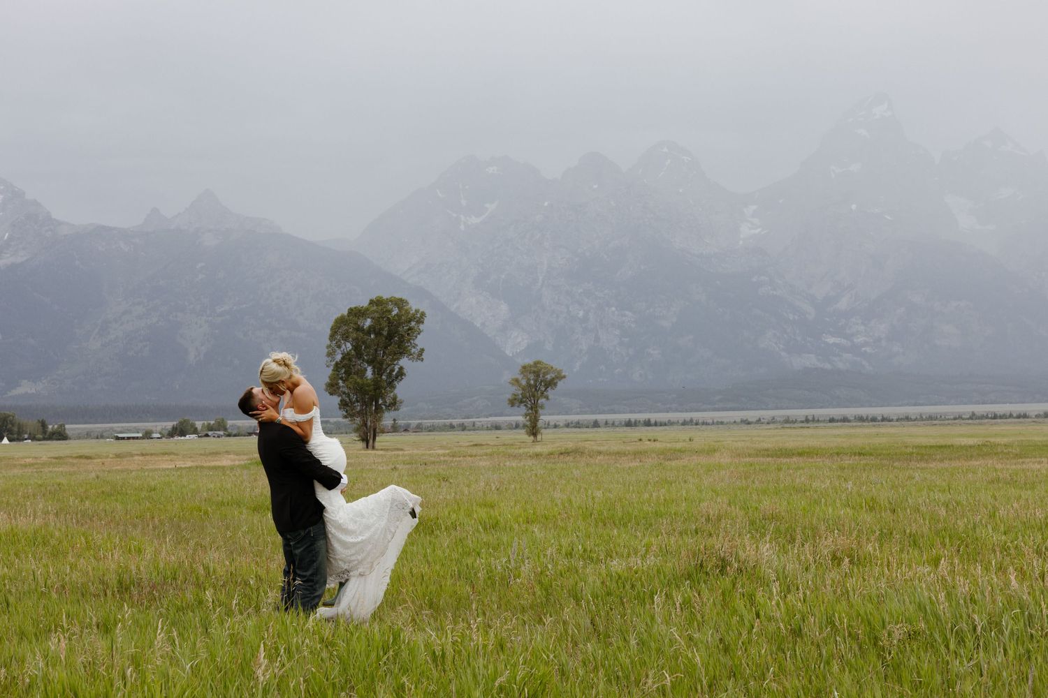 Bride and groom embrace in a vast green meadow with misty Grand Teton mountains in the background.