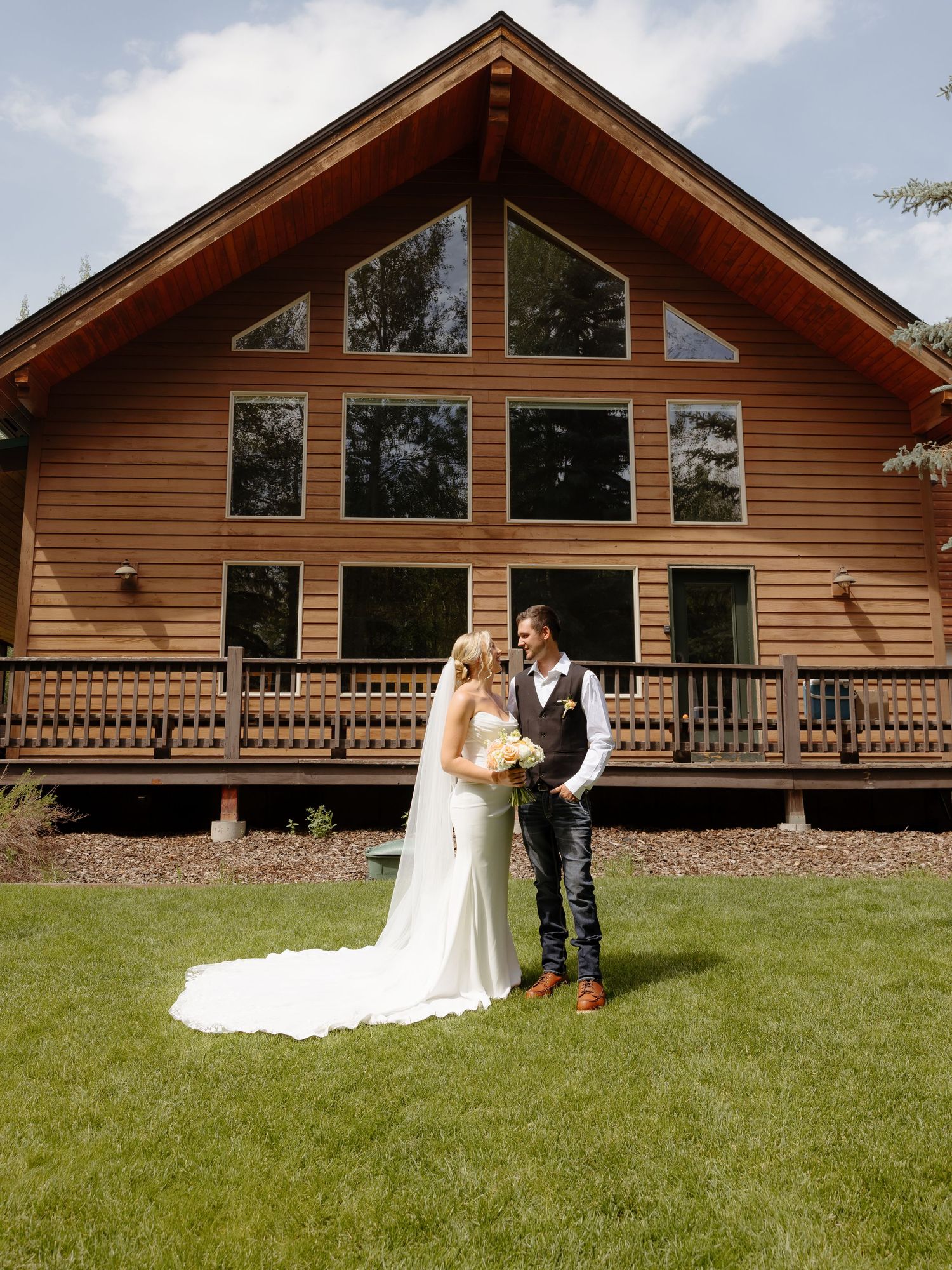 Bride in cathedral veil and groom in vest share a moment on green lawn before rustic log cabin venue.