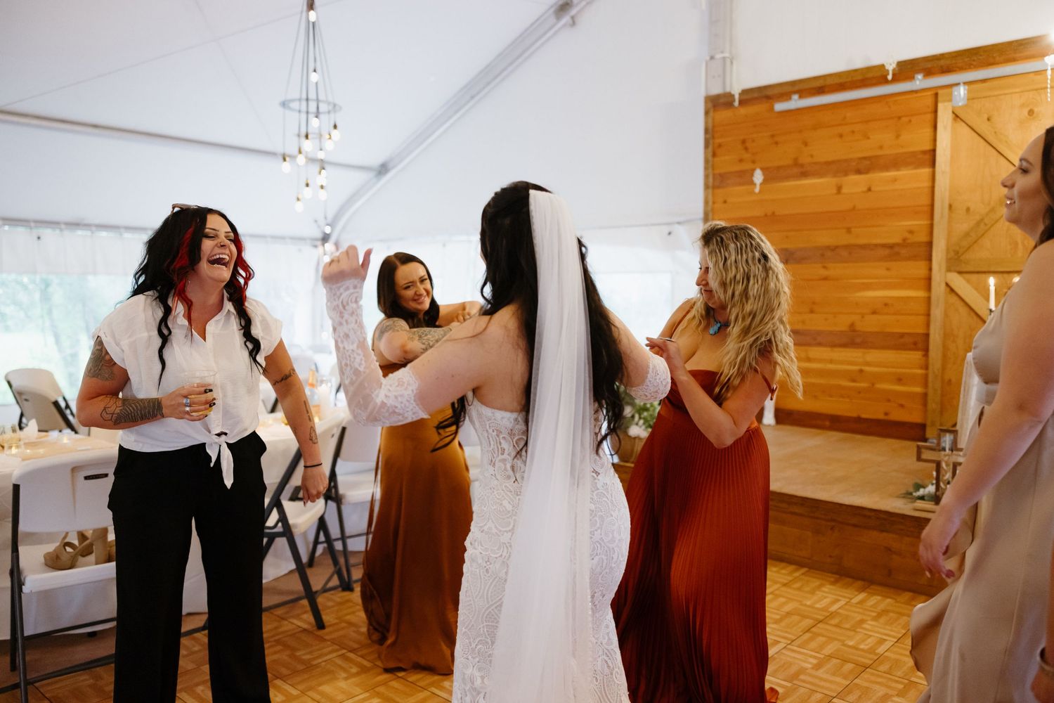 Bride celebrating with bridesmaids in rust and burgundy dresses at rustic tented wedding reception.