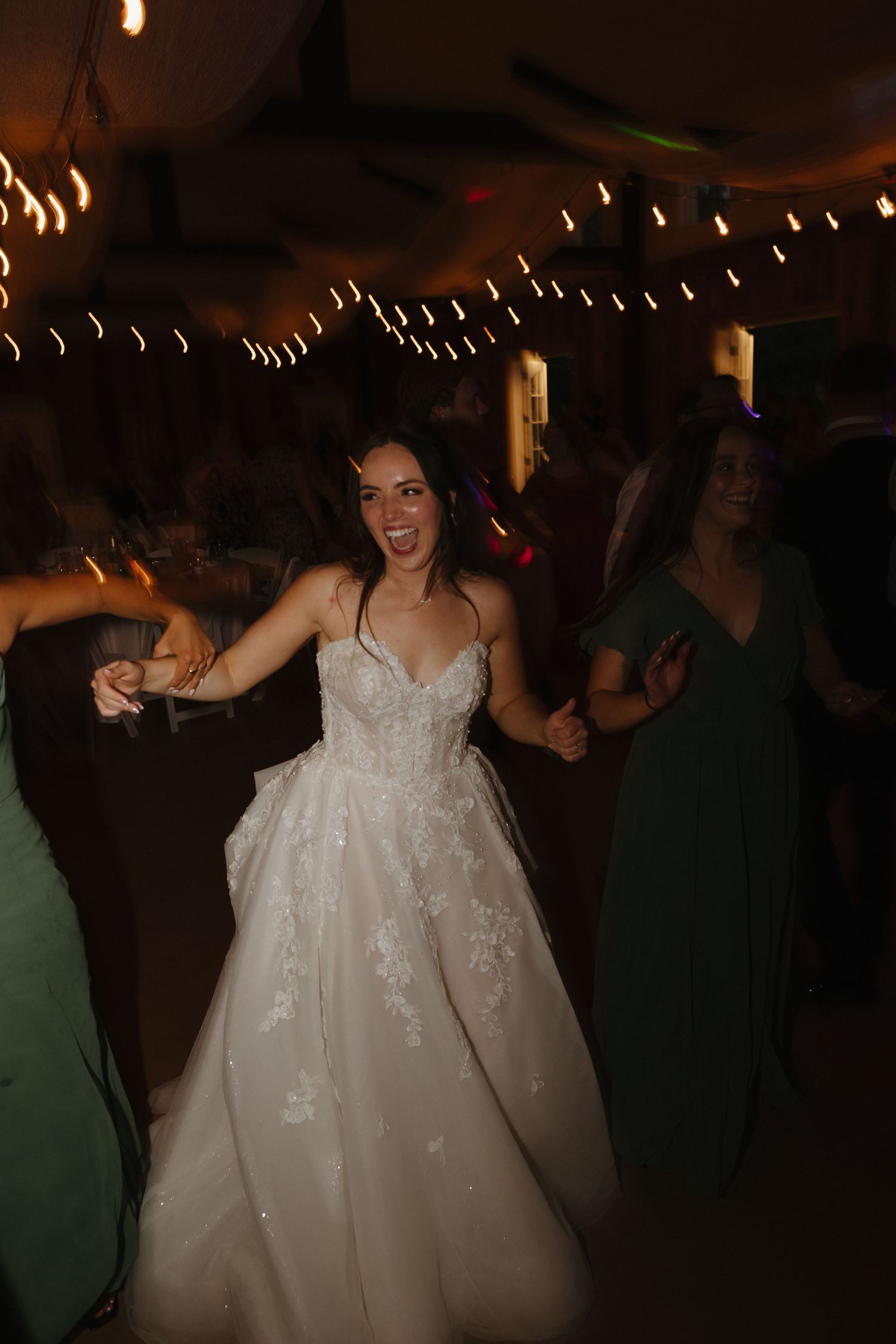 Smiling bride in lace ball gown dances joyfully with guests at wedding reception.