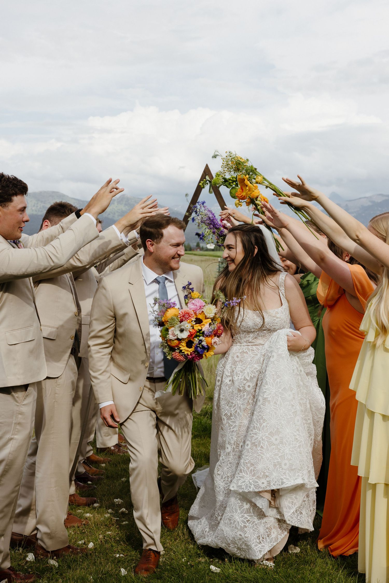 Bride and groom walk through arch of raised hands formed by wedding party on a scenic mountain backdrop.