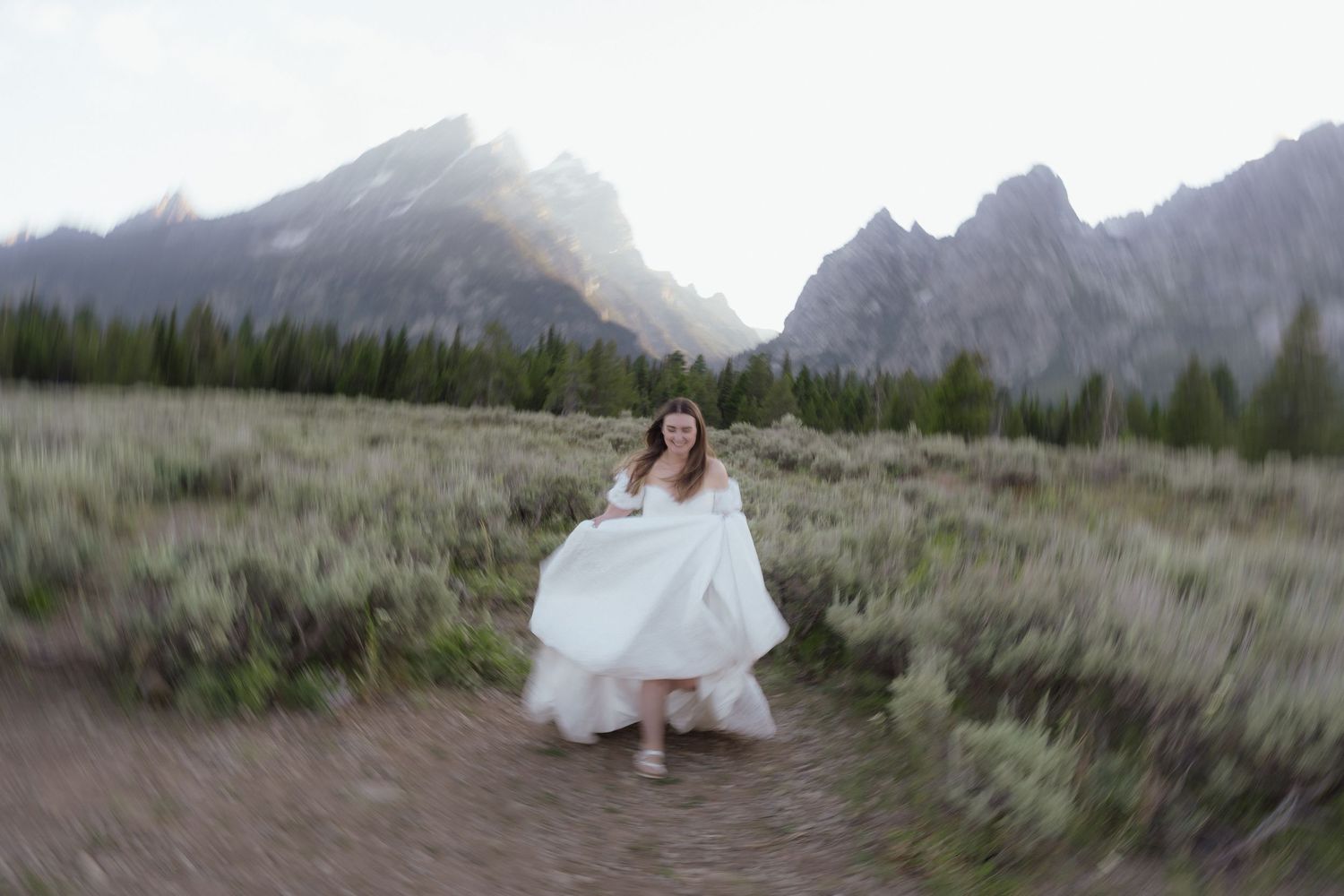 Bride in white dress runs playfully along a path through sagebrush with misty Grand Teton mountains behind.