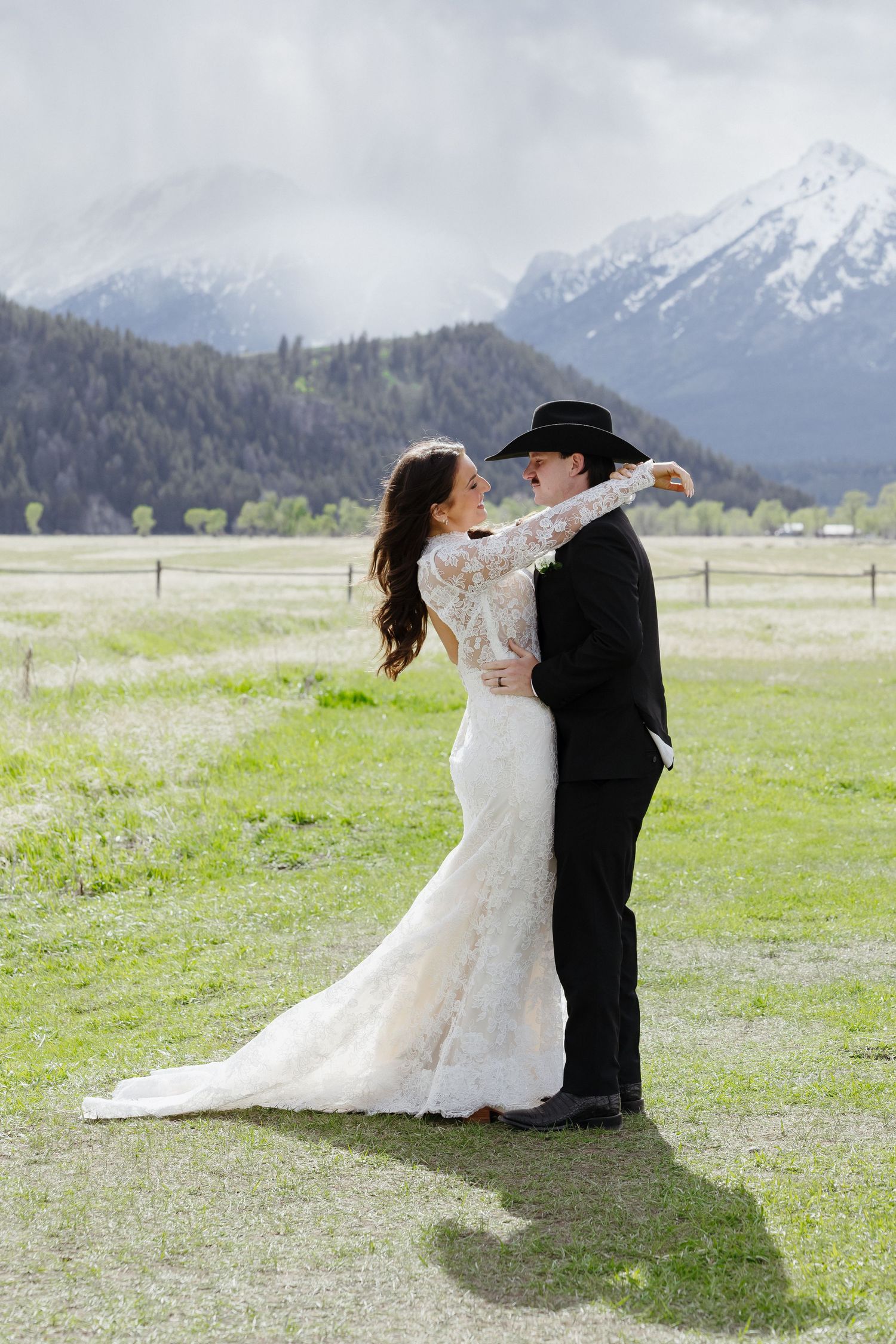 Western wedding couple embrace in green meadow with dramatic snow-capped mountain range in the background.