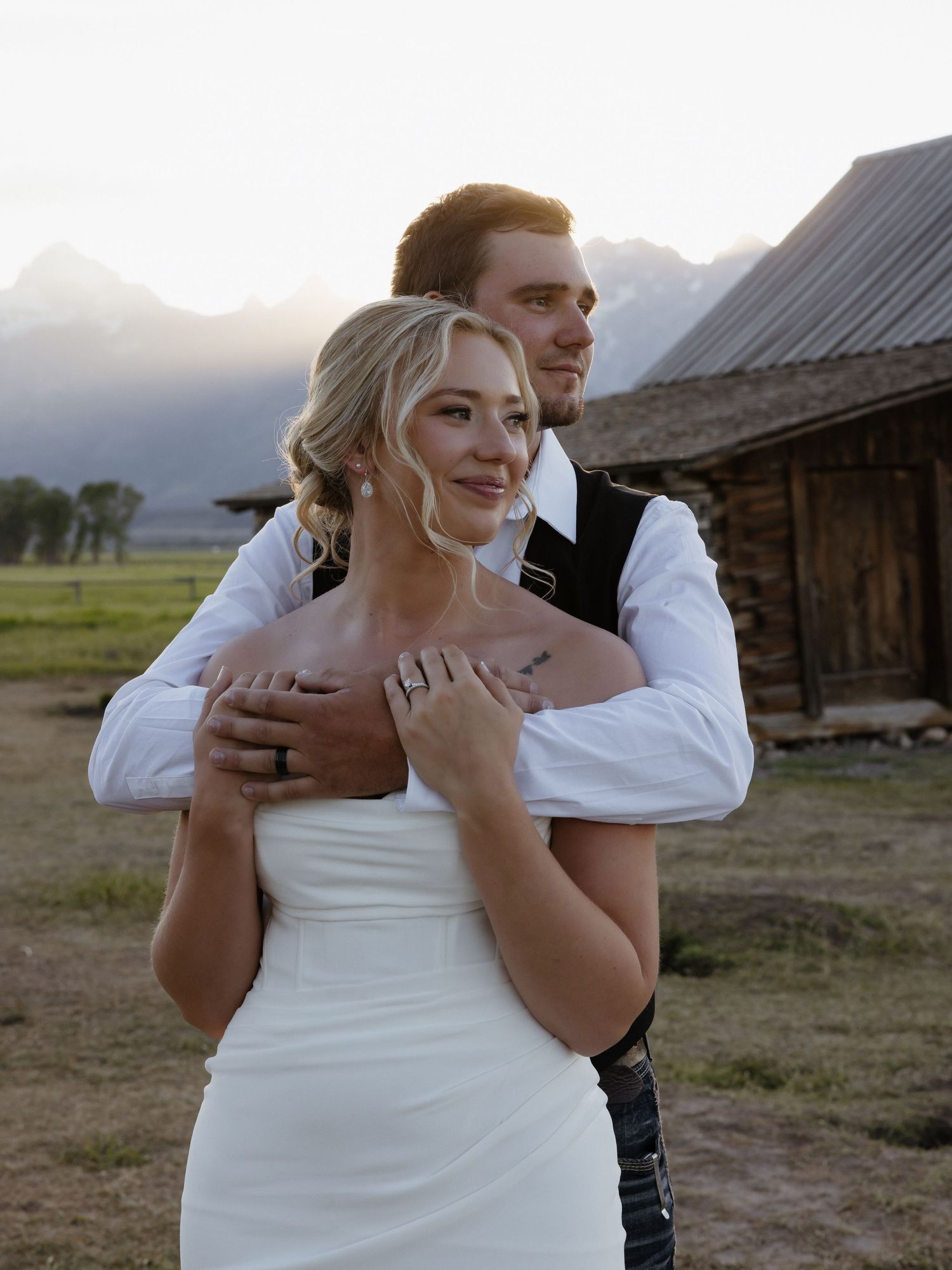 Groom tenderly embraces bride in white strapless gown near historic log cabin at sunset in Wyoming.
