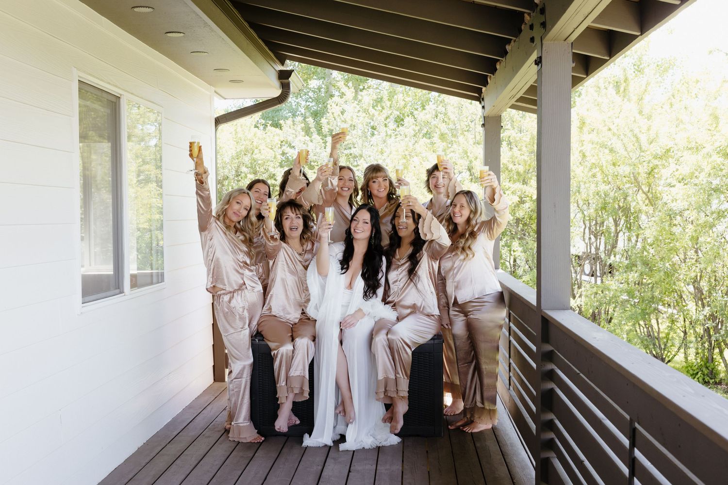 Bride and bridesmaids in satin robes celebrating on a porch, raising glasses in a joyful group photo.