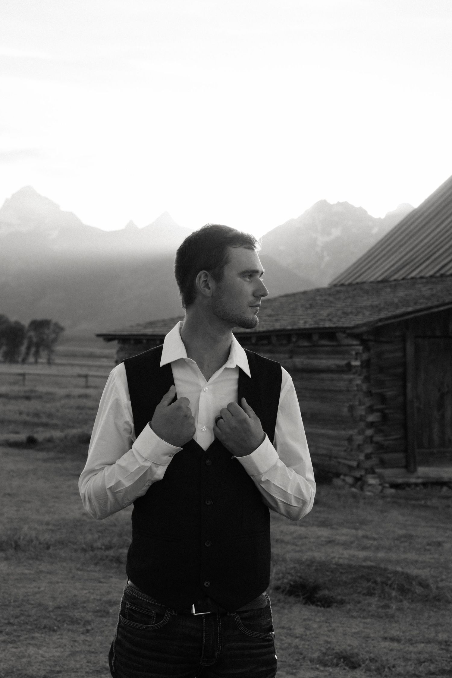 Groom in white shirt and black vest adjusting collar with mountain backdrop in black and white wedding photo.
