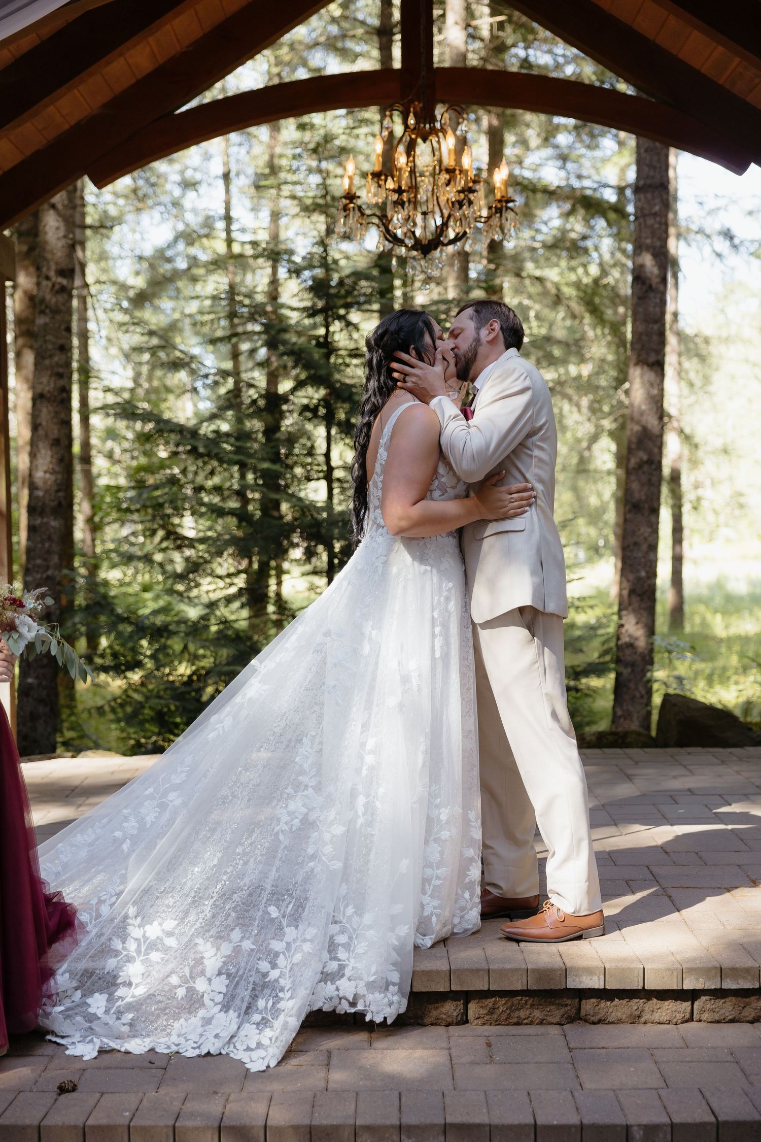 Bride and groom share first kiss under chandelier gazebo surrounded by tall pine trees at outdoor forest wedding.