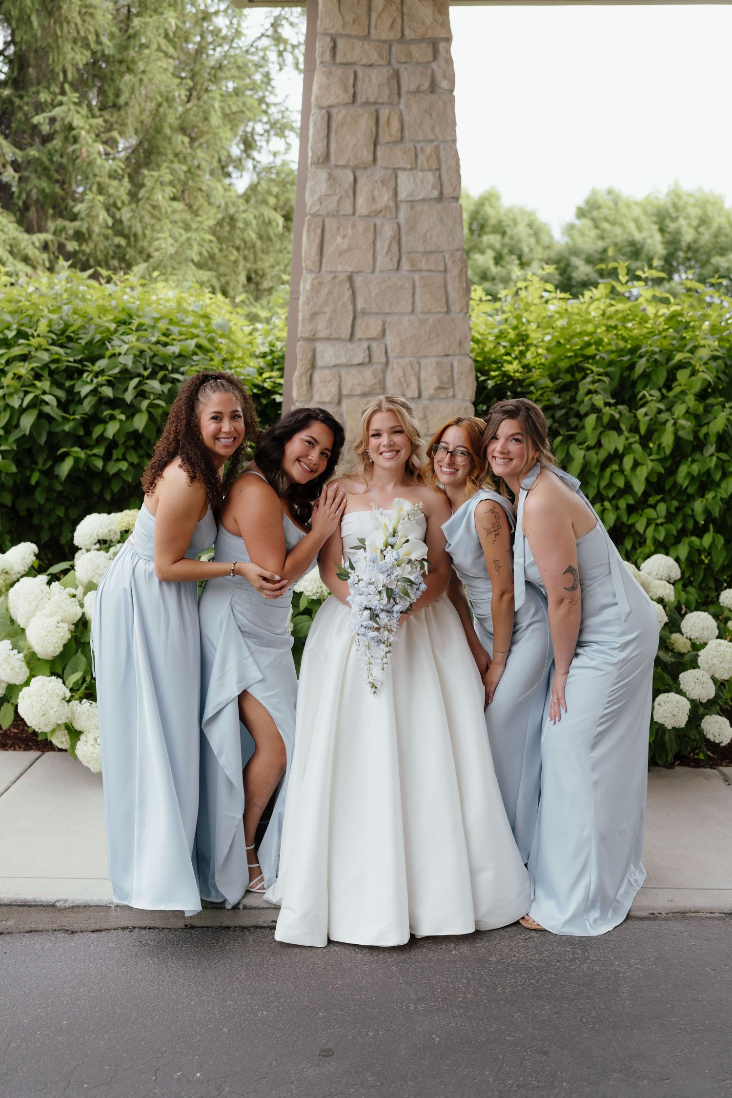 Bride in white gown holds bouquet surrounded by four bridesmaids in light blue dresses near white hydrangeas.