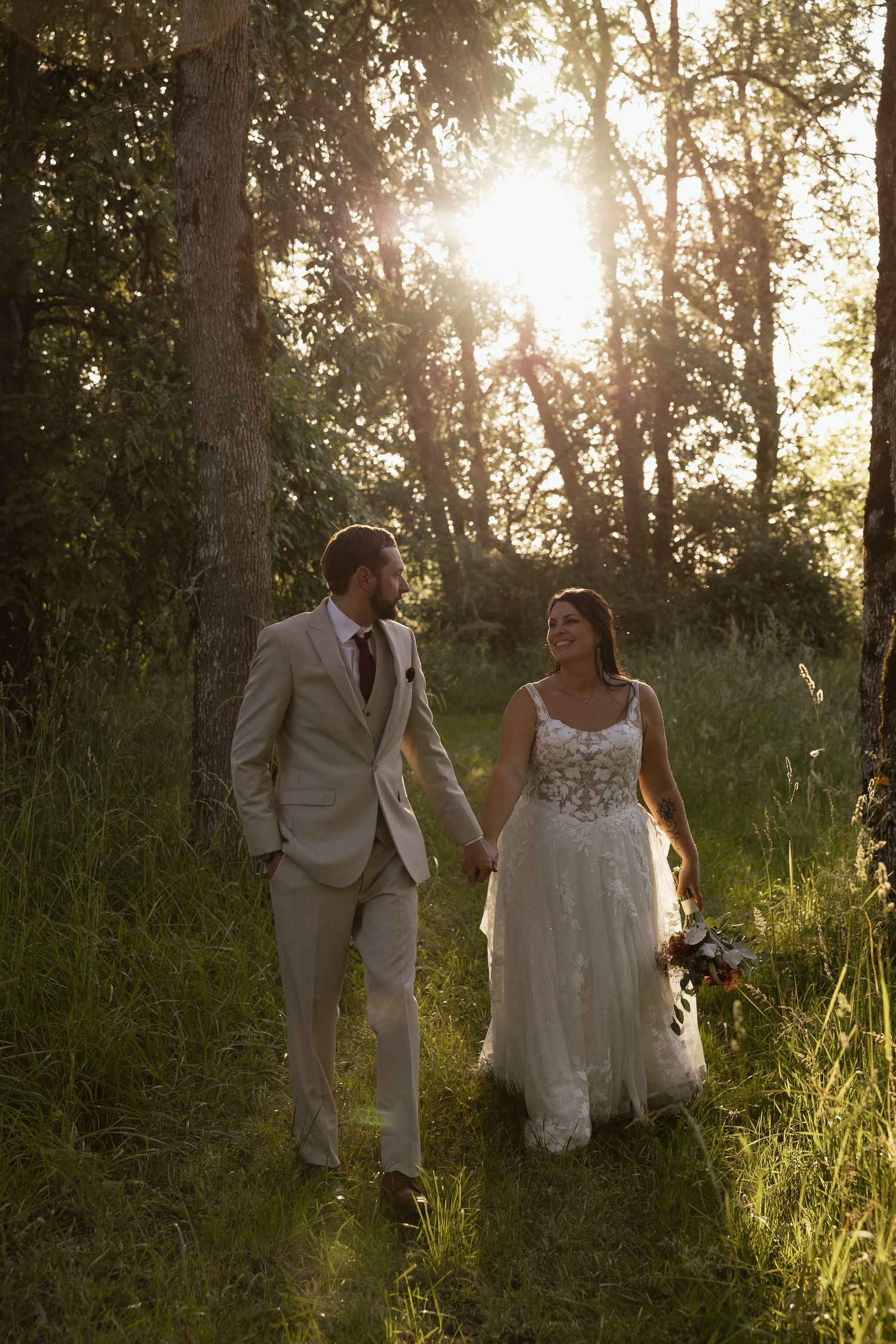 Bride and groom holding hands walking through golden sunlit forest meadow during romantic sunset wedding portraits.