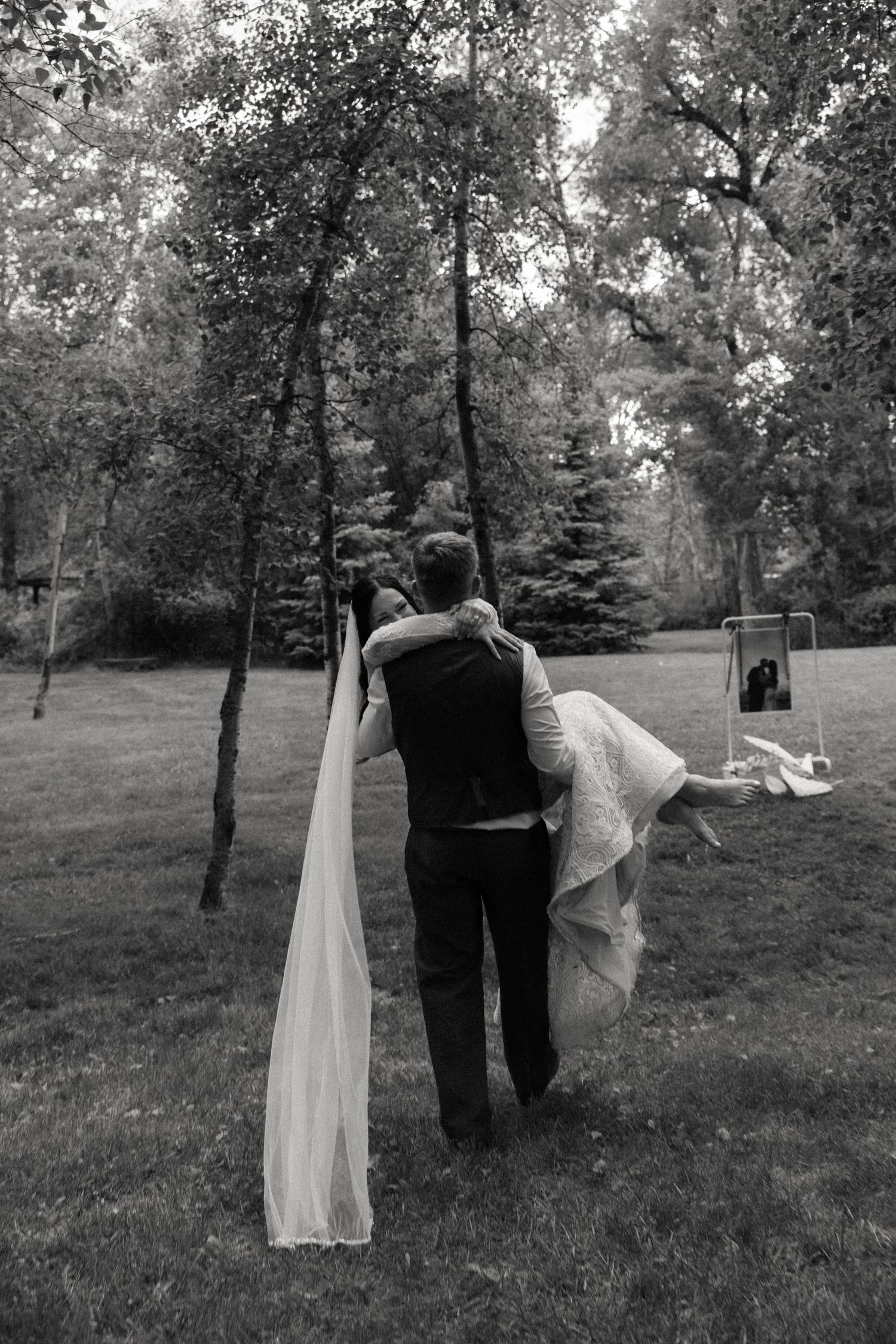 Black and white photo of groom carrying bride through a wooded garden at their wedding.