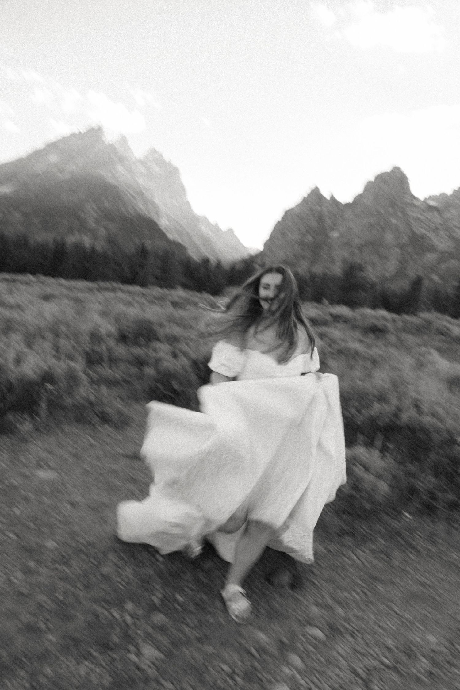 Black and white blurred artistic photo of bride joyfully running in white gown with mountain backdrop.