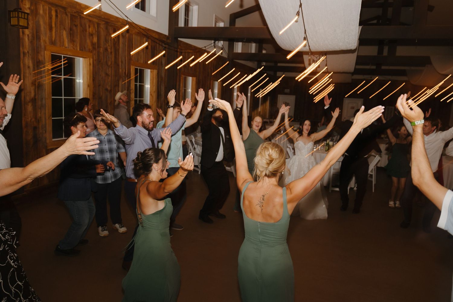 Wedding guests celebrate on dance floor with hands raised in rustic barn reception hall.