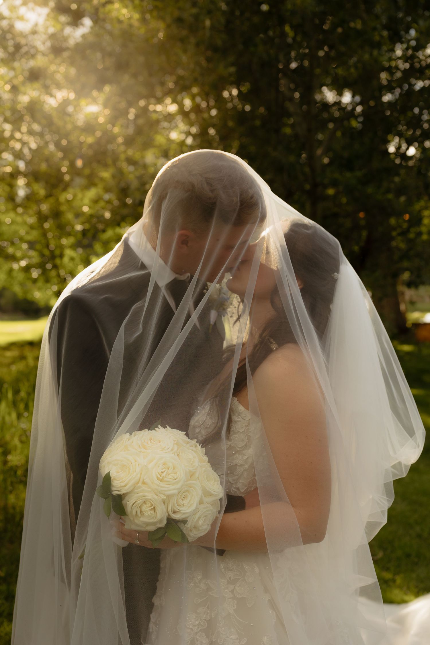 Bride and groom kissing under flowing white veil in golden sunlight holding white rose bouquet.