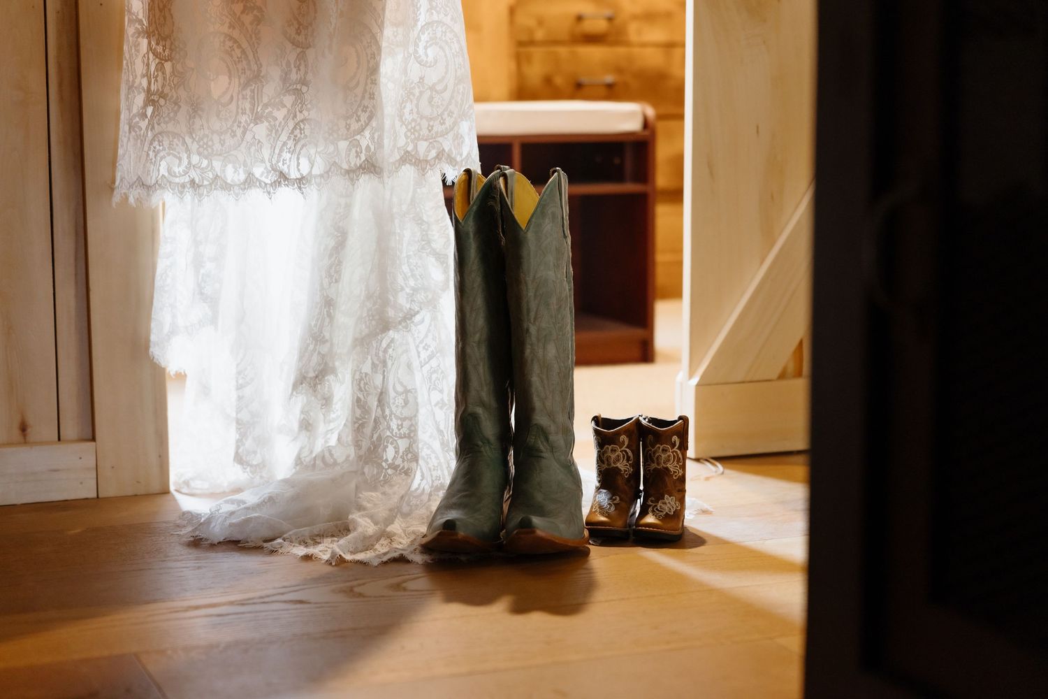 Turquoise cowboy boots and mini brown boots beside a lace wedding dress on a rustic wooden floor.