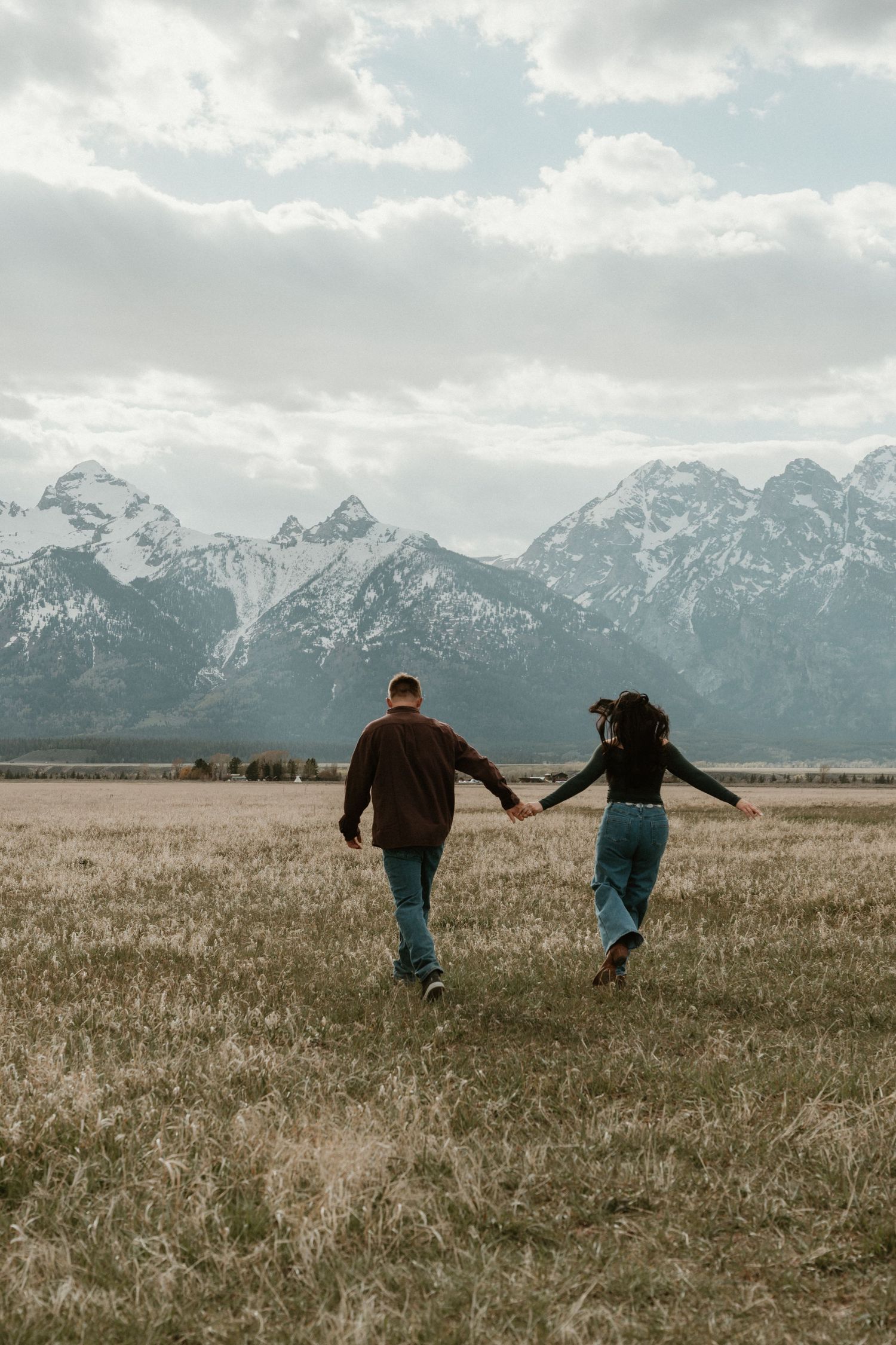 Couple holding hands while walking through a golden field toward the majestic Grand Teton mountains.