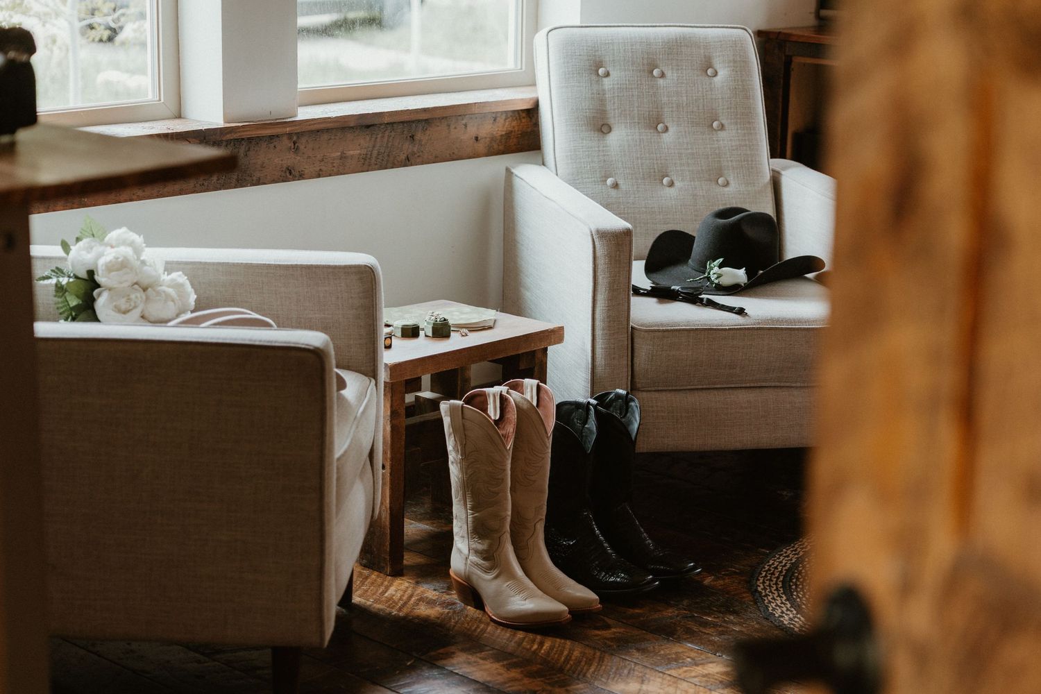 Cowboy boots and wedding accessories arranged near white tufted chairs on hardwood floor.