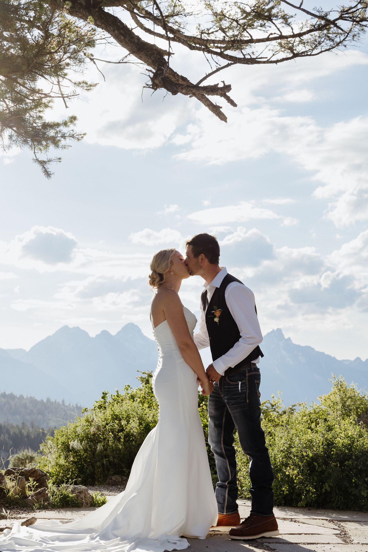 Bride and groom kissing while holding hands under pine tree with Grand Teton mountain views.