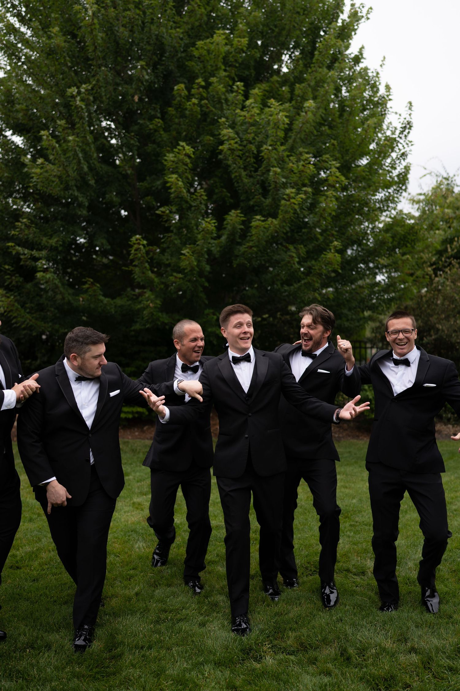 Six groomsmen in black tuxedos with bow ties laugh and pose together outdoors on green grass with trees behind them.