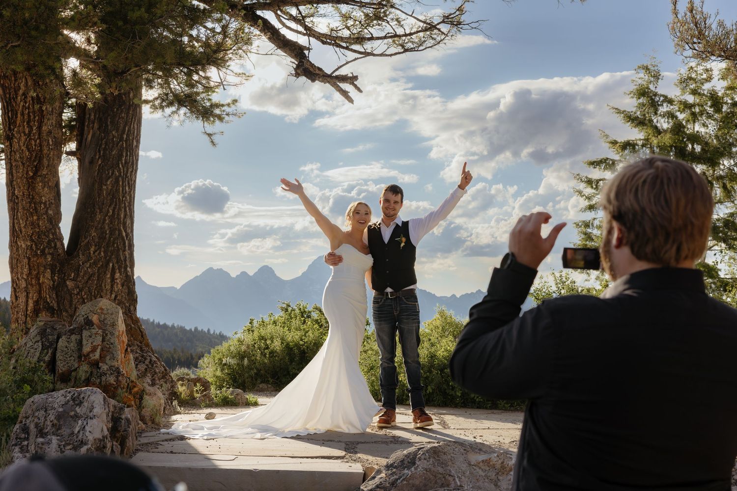 Photographer captures jubilant bride and groom raising arms in celebration on scenic overlook with Teton peaks behind.