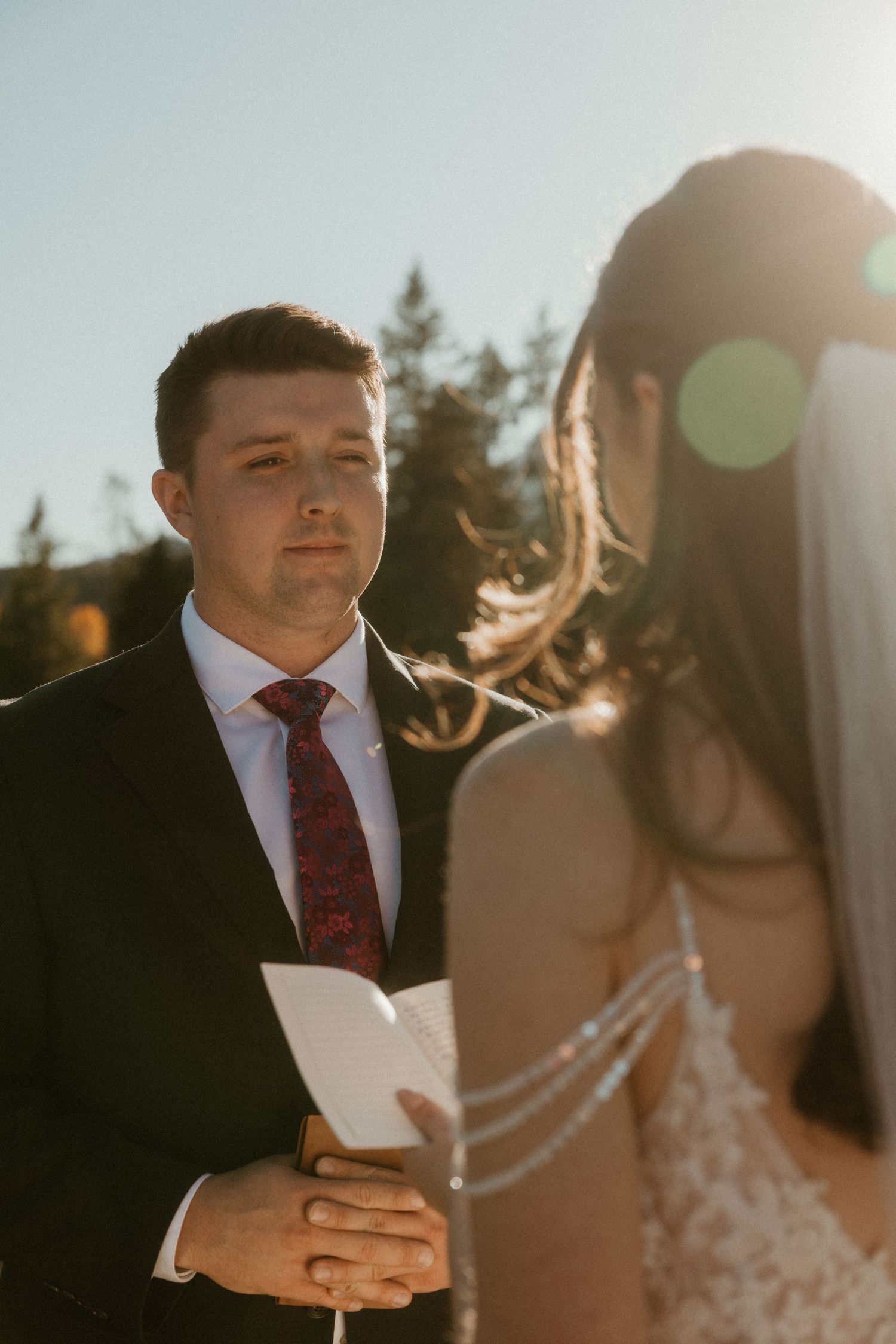 Groom reads wedding vows from paper while bride in lace dress and veil stands before him outdoors.