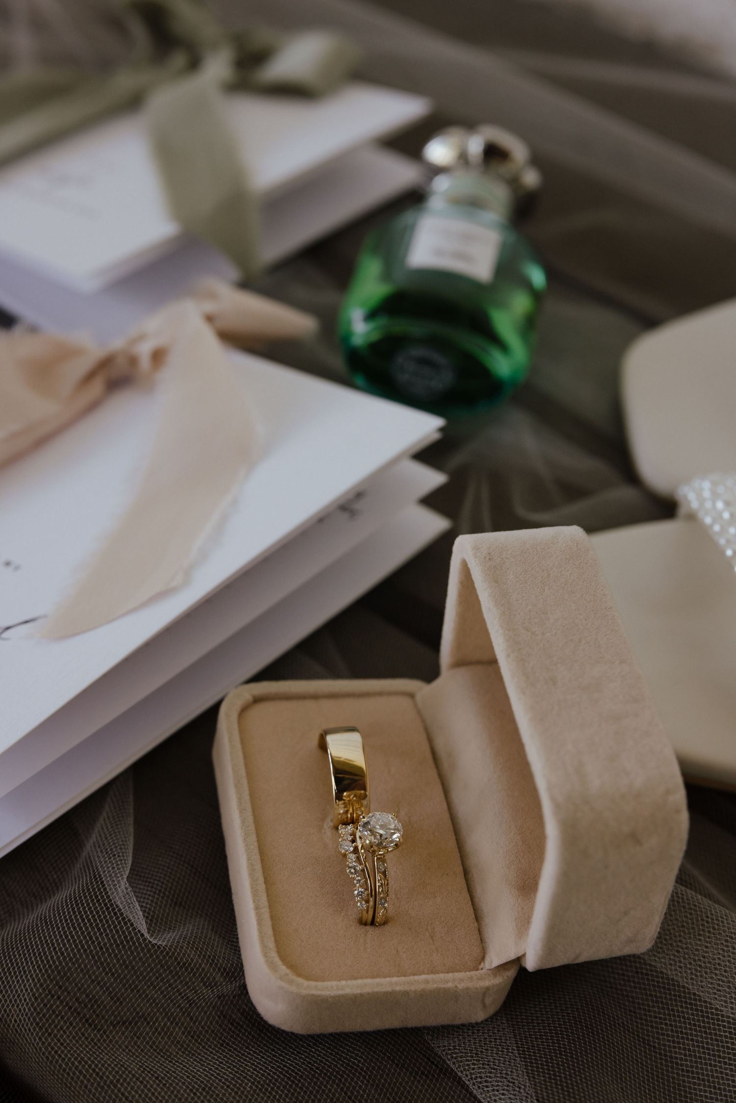 Gold diamond engagement ring displayed in velvet box beside vow books and green perfume bottle on fabric.