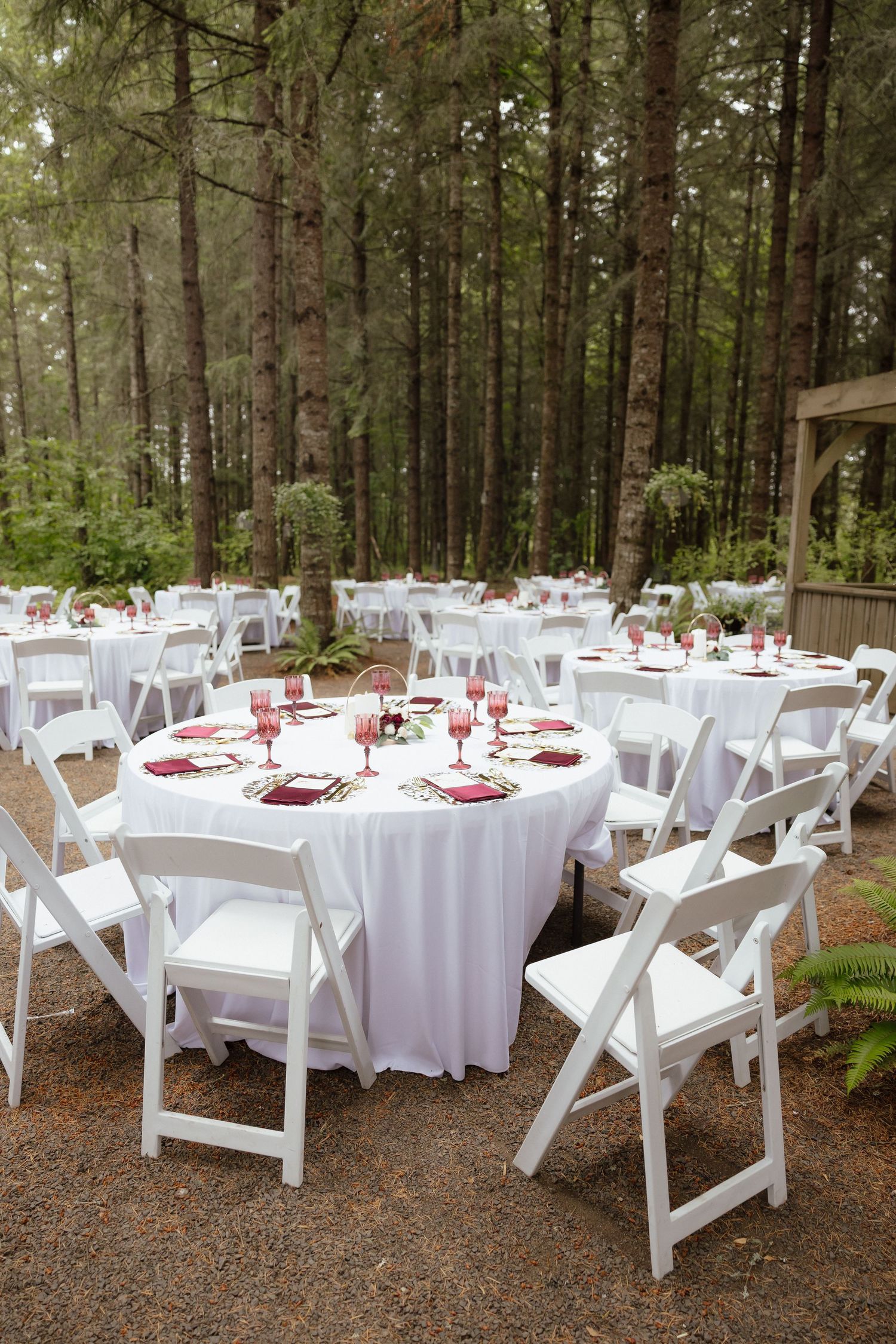 Outdoor forest wedding reception with white round tables, white folding chairs, and burgundy napkins set among tall pine trees.