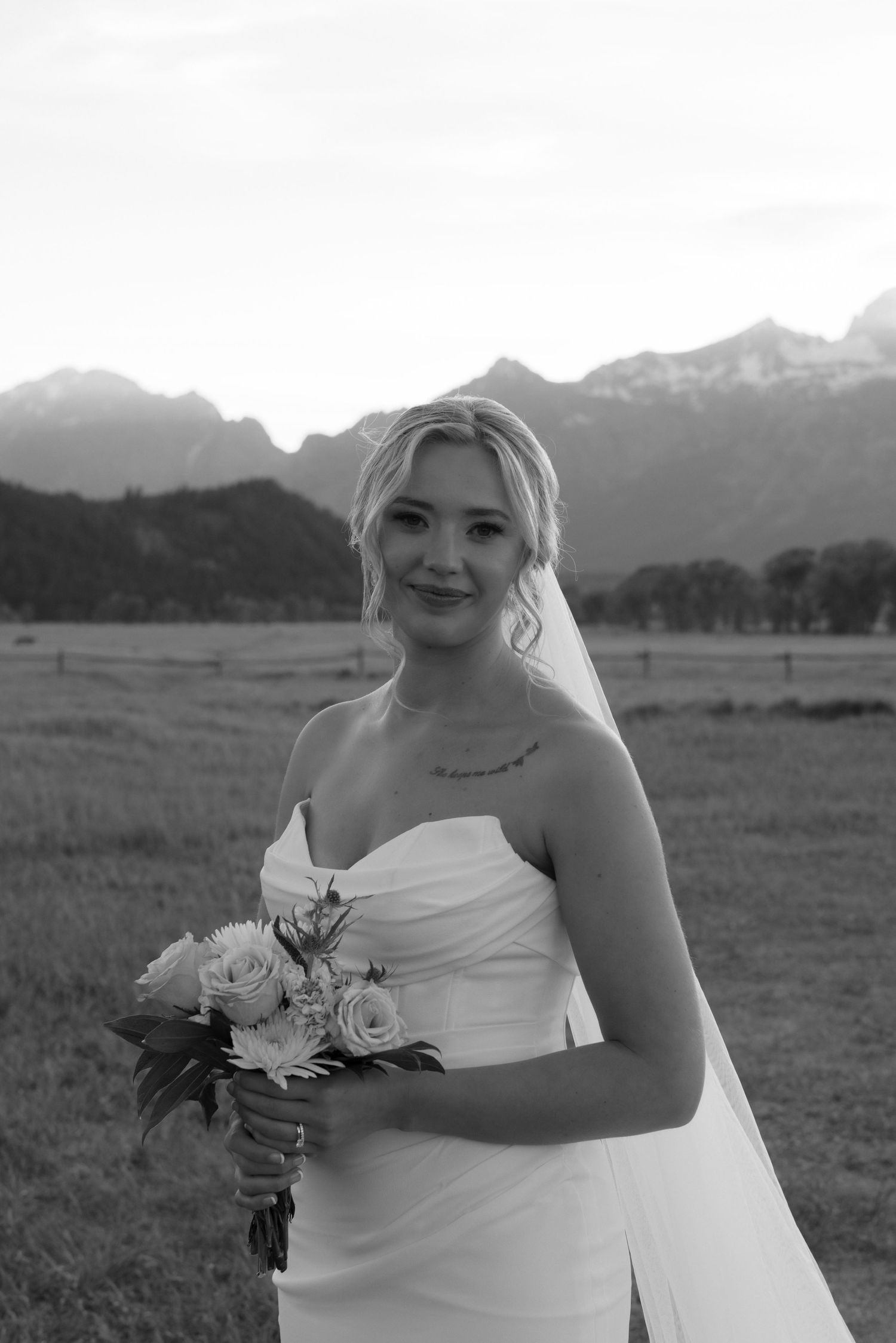 Black and white portrait of smiling blonde bride holding bouquet with Grand Teton mountains in background.