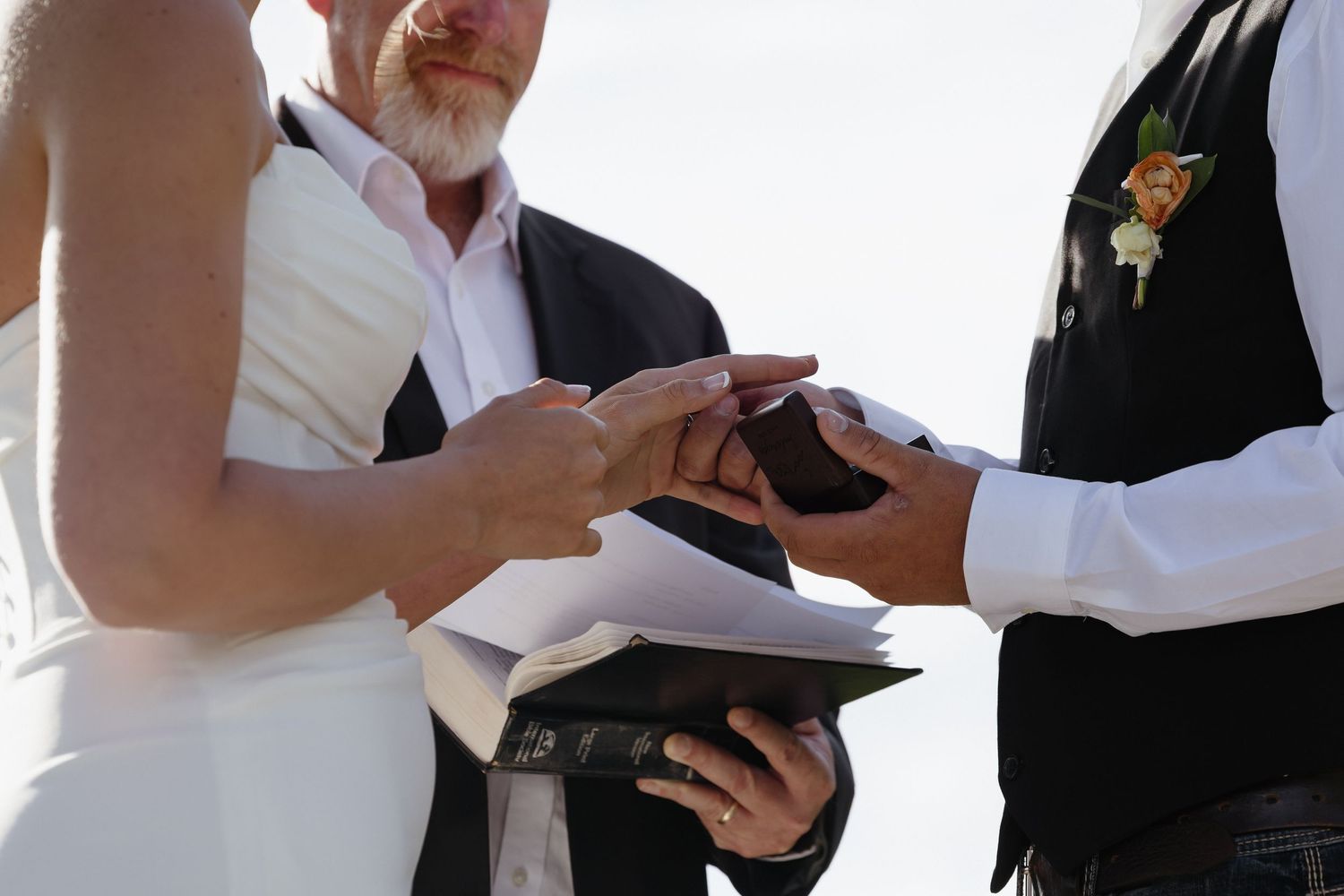 Officiant placing wedding ring on bride's finger during outdoor ceremony with open Bible.