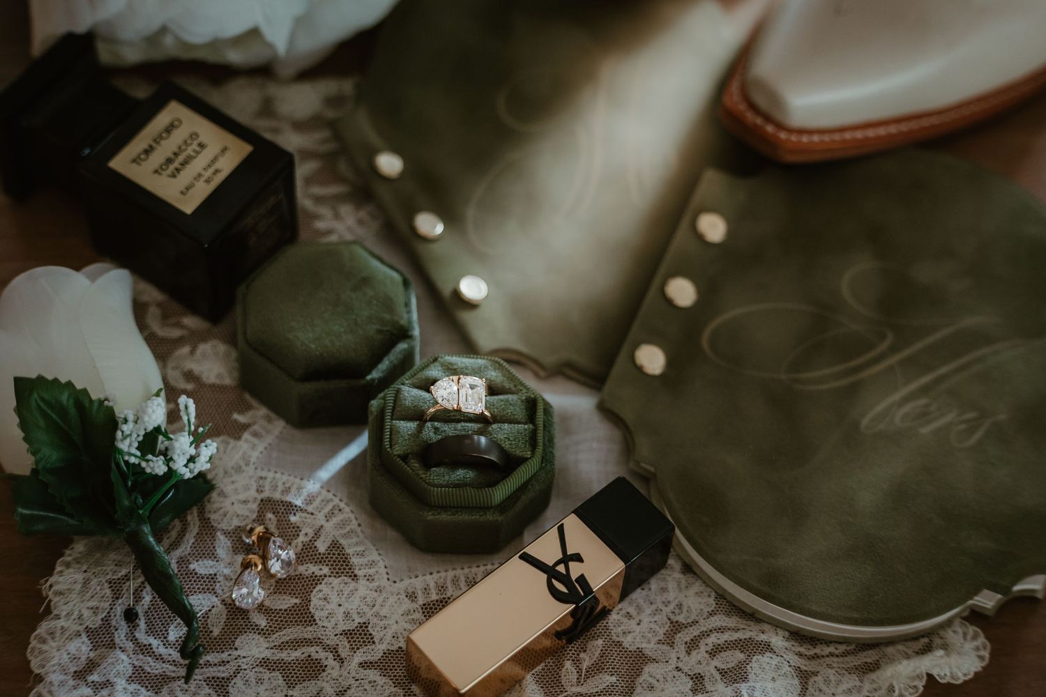Green velvet ring box with wedding rings, YSL perfume, and leather goods on lace fabric.