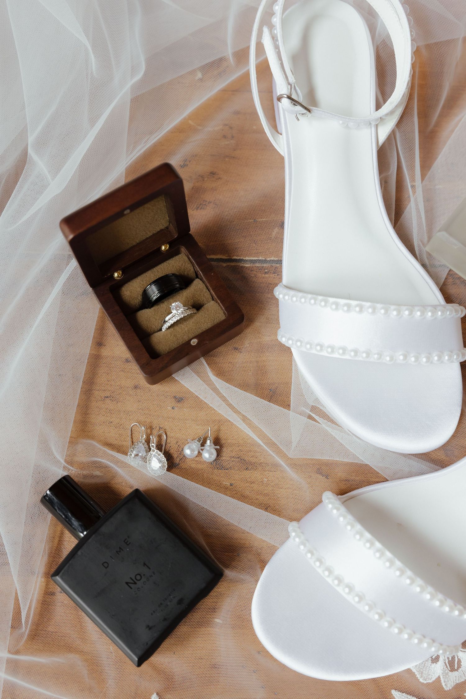 Flat lay of white pearl bridal heels, wedding rings in wooden box, pearl earrings, and black perfume bottle.