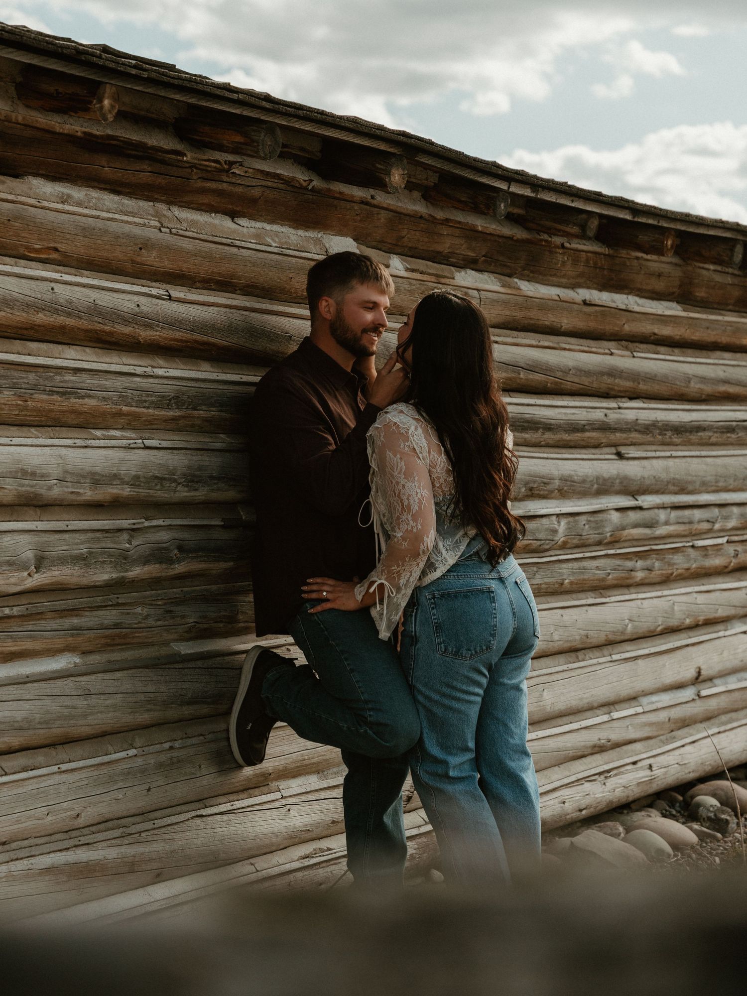 Couple leaning against a rustic wooden wall, smiling and about to kiss during an outdoor photo session.