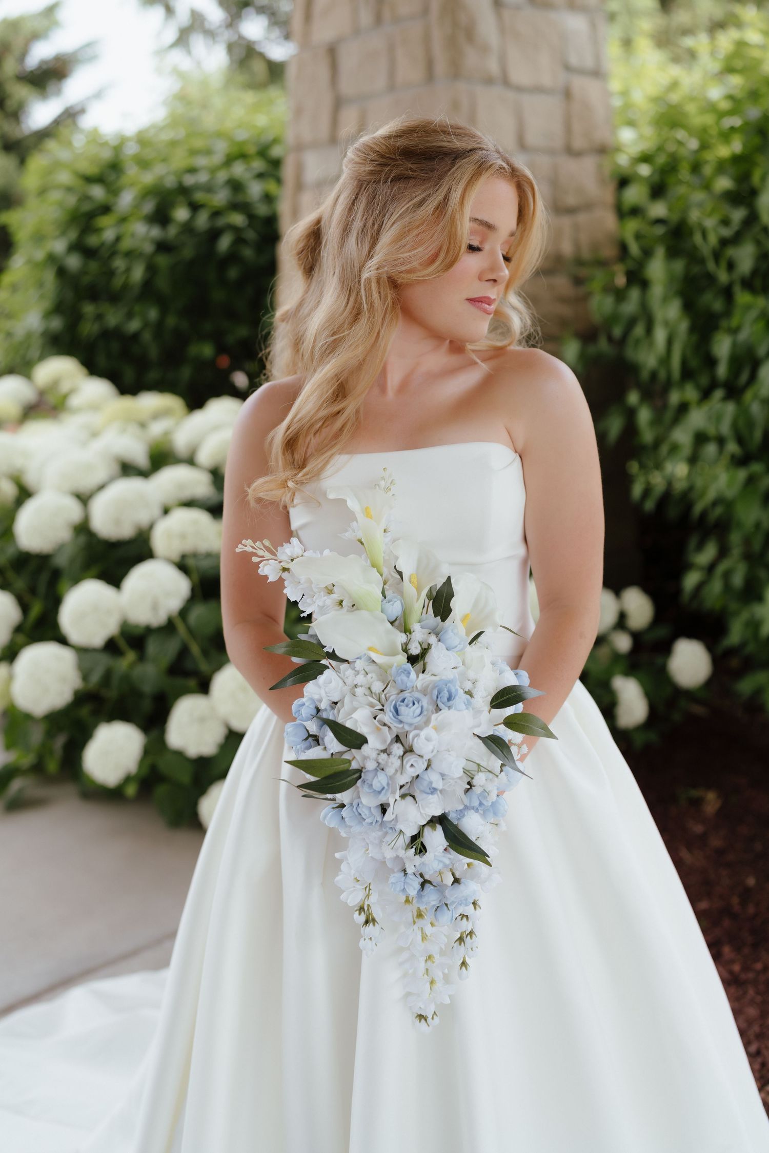Bride in strapless white ball gown holds cascading blue and white floral bouquet outdoors near hydrangeas.