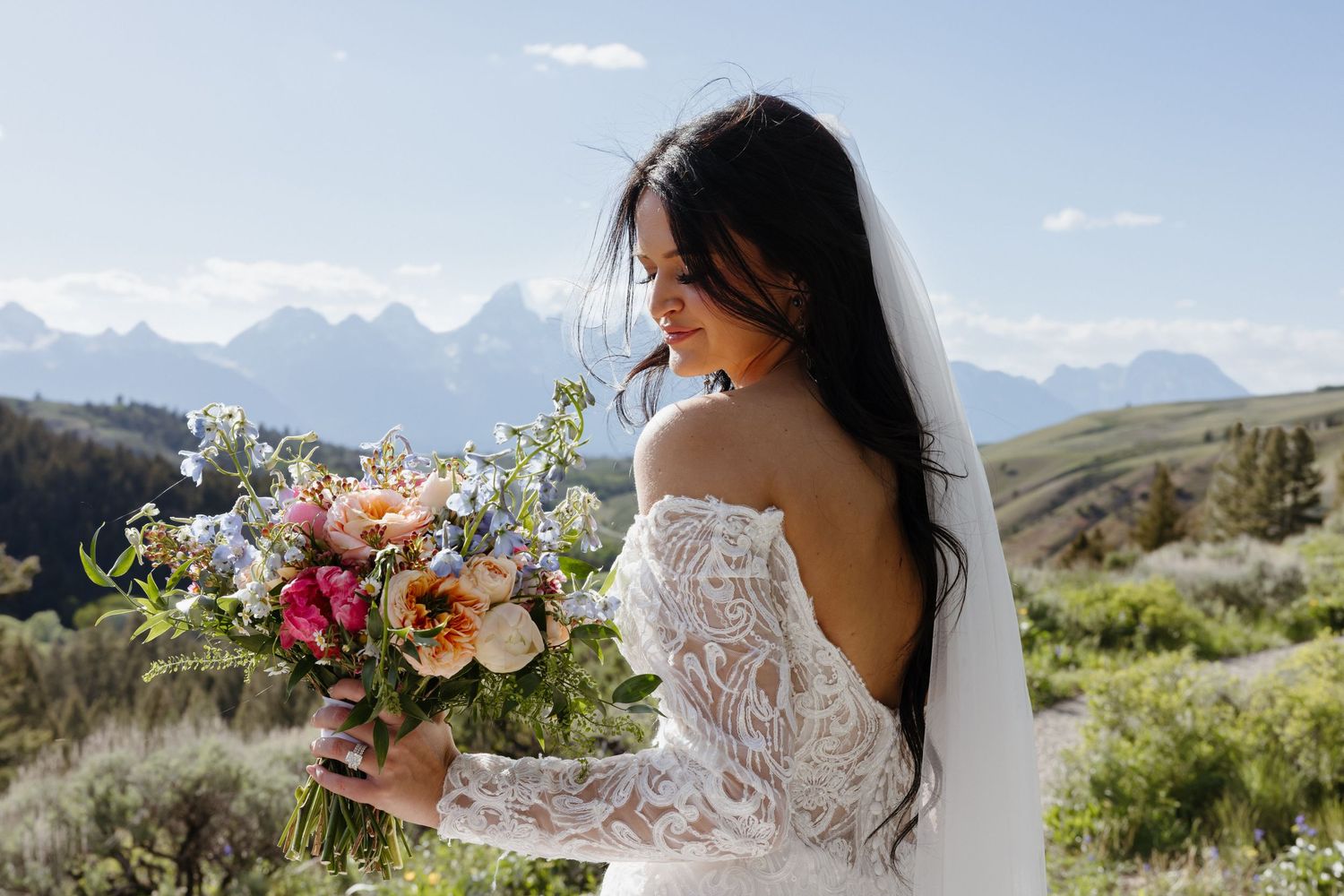 Bride in off-shoulder lace gown admires her colorful wildflower bouquet against a mountain landscape.