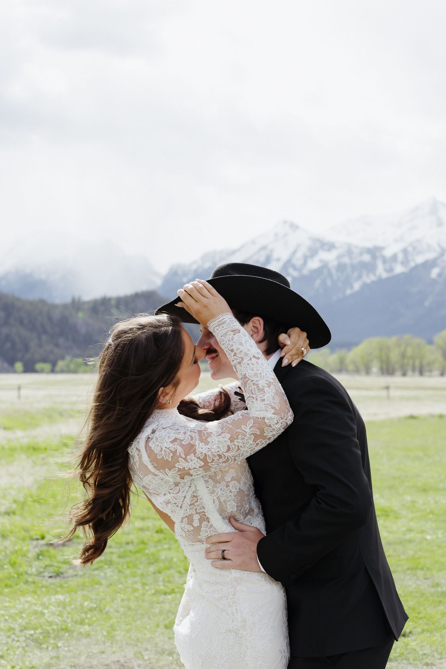 Bride and groom share a kiss under black cowboy hat with Grand Teton snow-capped mountains in background.