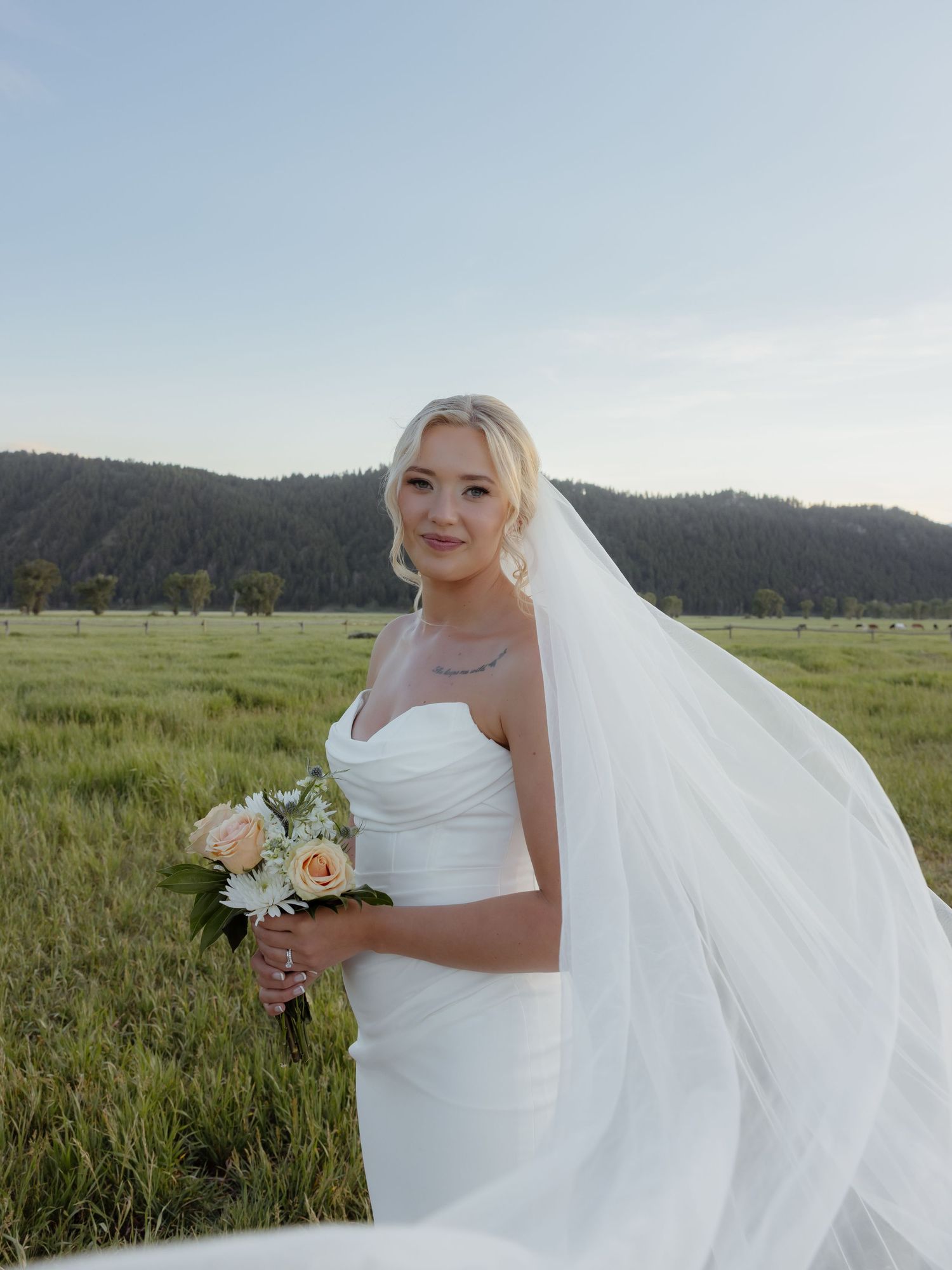 Radiant bride in white strapless gown holding bouquet with flowing veil in green mountain meadow.