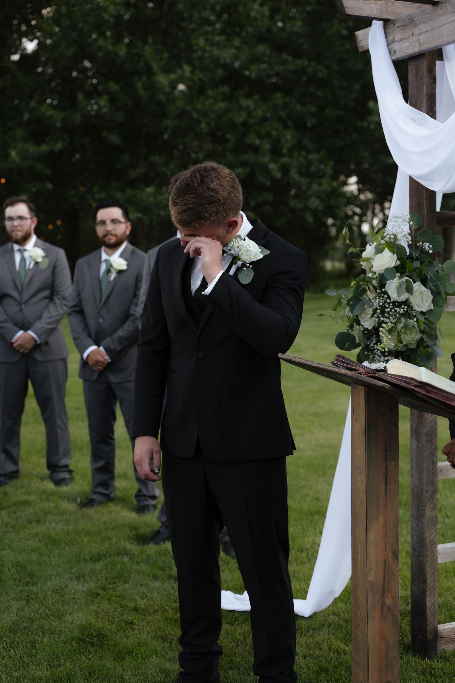 Emotional groom wipes tears at outdoor wedding altar with groomsmen standing behind.