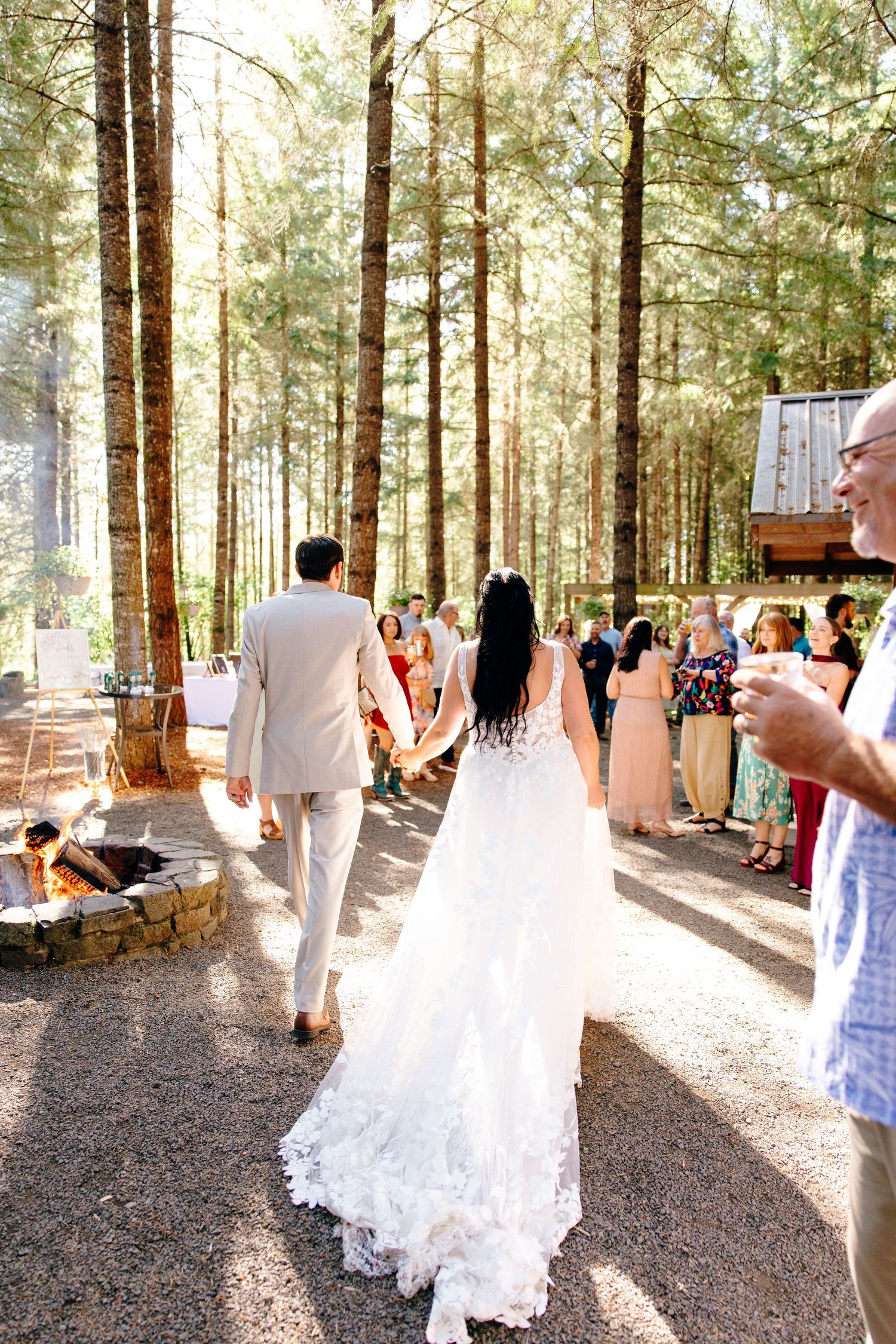Bride in lace gown and groom in tan suit walk hand-in-hand through pine forest as guests cheer at reception.