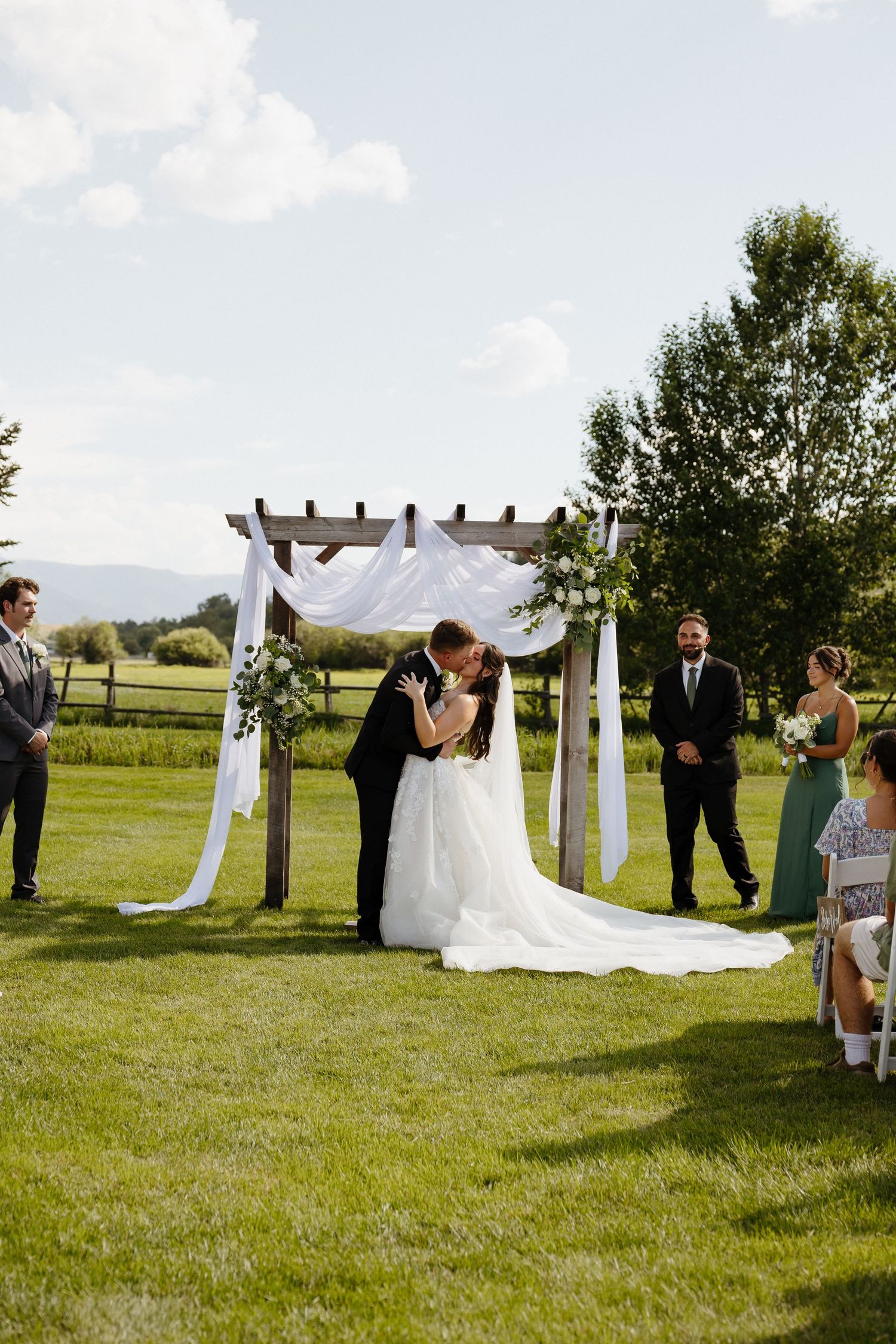 Bride and groom share first kiss under rustic wooden arch with white draping outdoors.