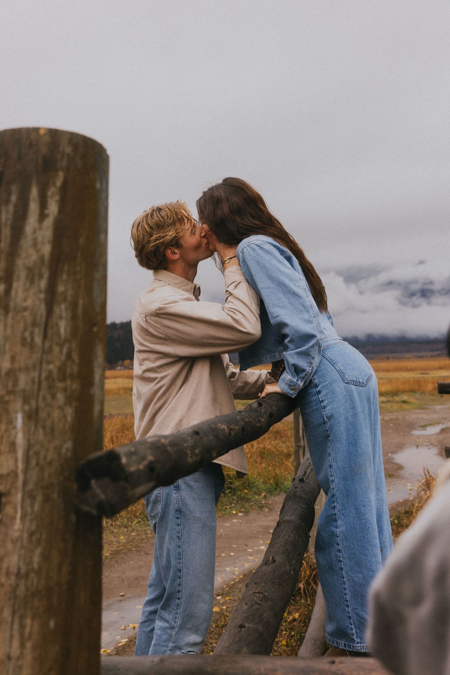 Couple sharing a romantic kiss by a rustic wooden fence post in an open field with misty mountains.