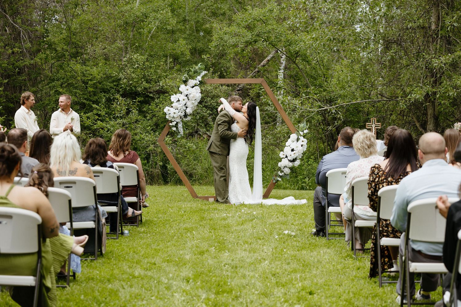 Outdoor wedding ceremony with couple kissing under hexagonal arch adorned with white flowers, surrounded by seated guests.