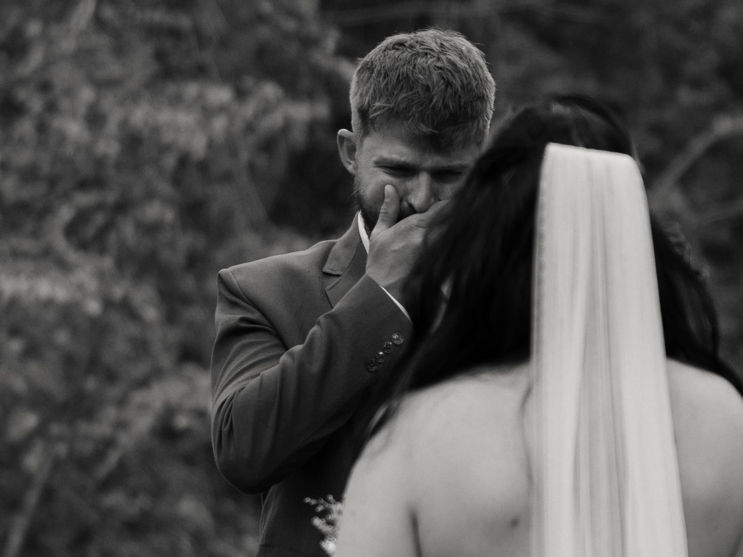Emotional black and white photo of groom covering his mouth in tears seeing bride for the first time.