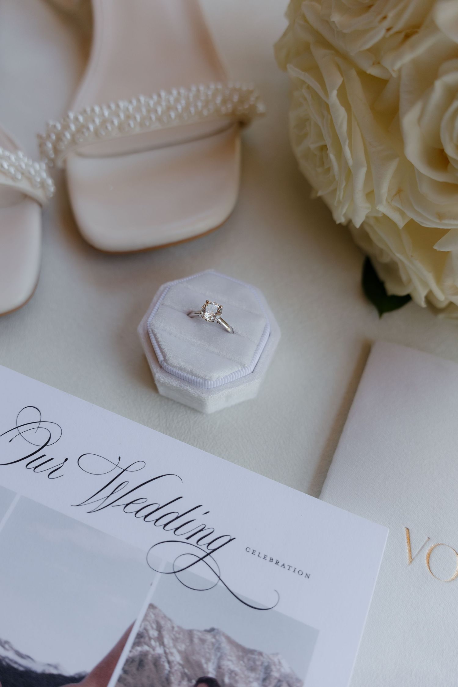 Elegant wedding flat lay with diamond ring in velvet box, pearl heels, white roses, and wedding program.