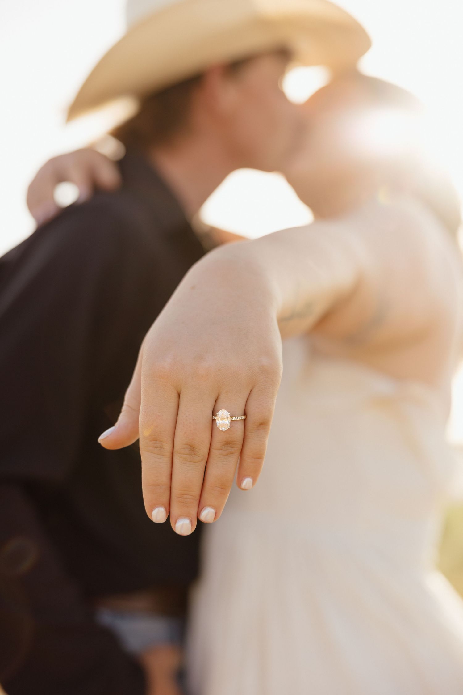 Close-up of an oval engagement ring on a woman's hand as couple kisses in golden sunlight.