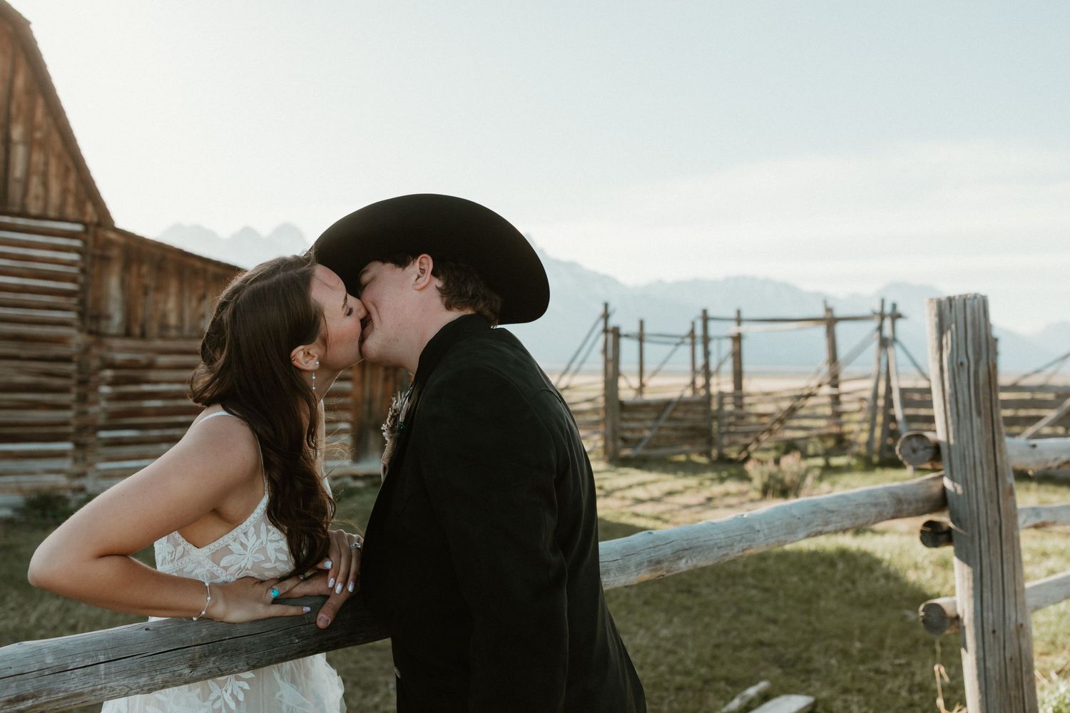 Bride and groom share a kiss leaning over wooden fence with rustic barn and mountains behind.